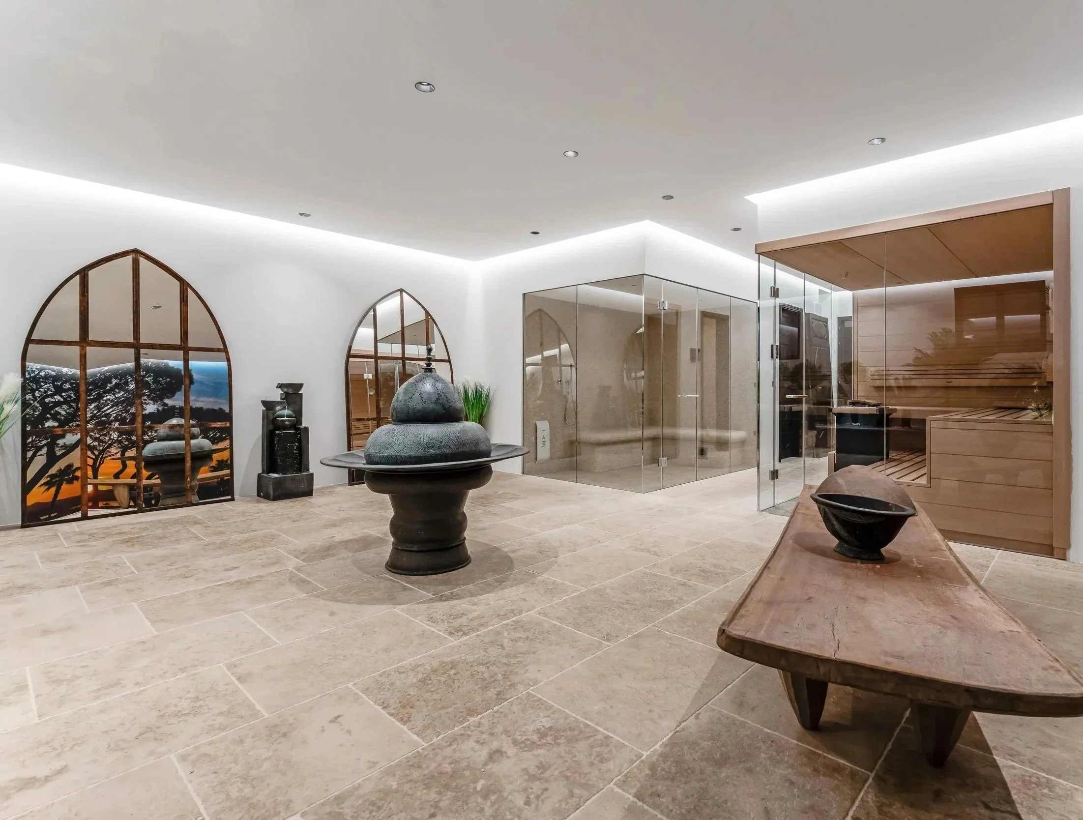 Modern spa room with beige tiled floor, white walls, and ceiling. Features arched windows with scenic sunset view, a black water fountain, and decorative statues. There is a glass sauna enclosure with wooden benches and a black heater, along with wooden accents and indoor plants.