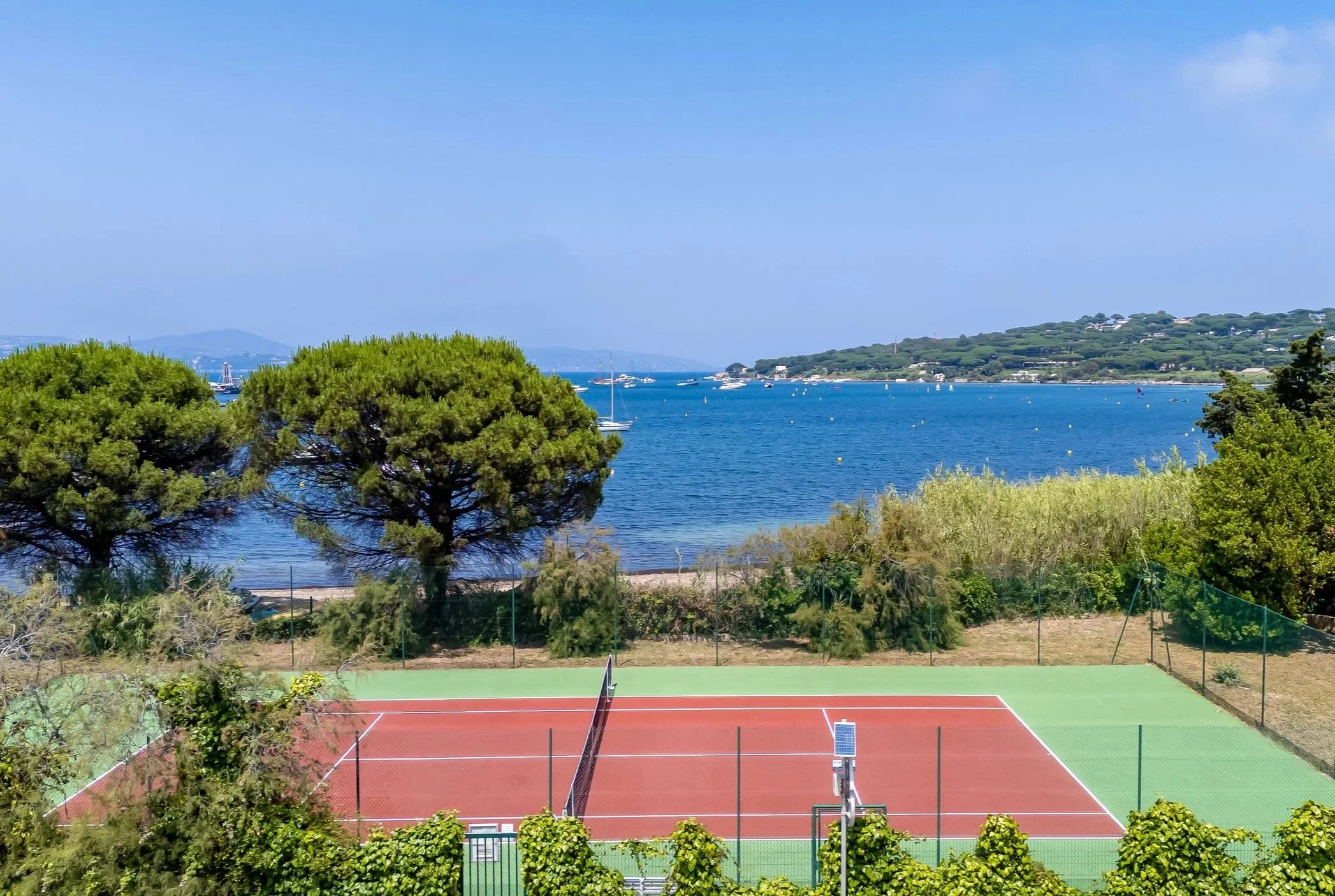 A red and green tennis court surrounded by a black fence, with pine trees and shrubbery, overlooking a blue lake with sailboats and a hilly landscape in the background on a clear, sunny day.