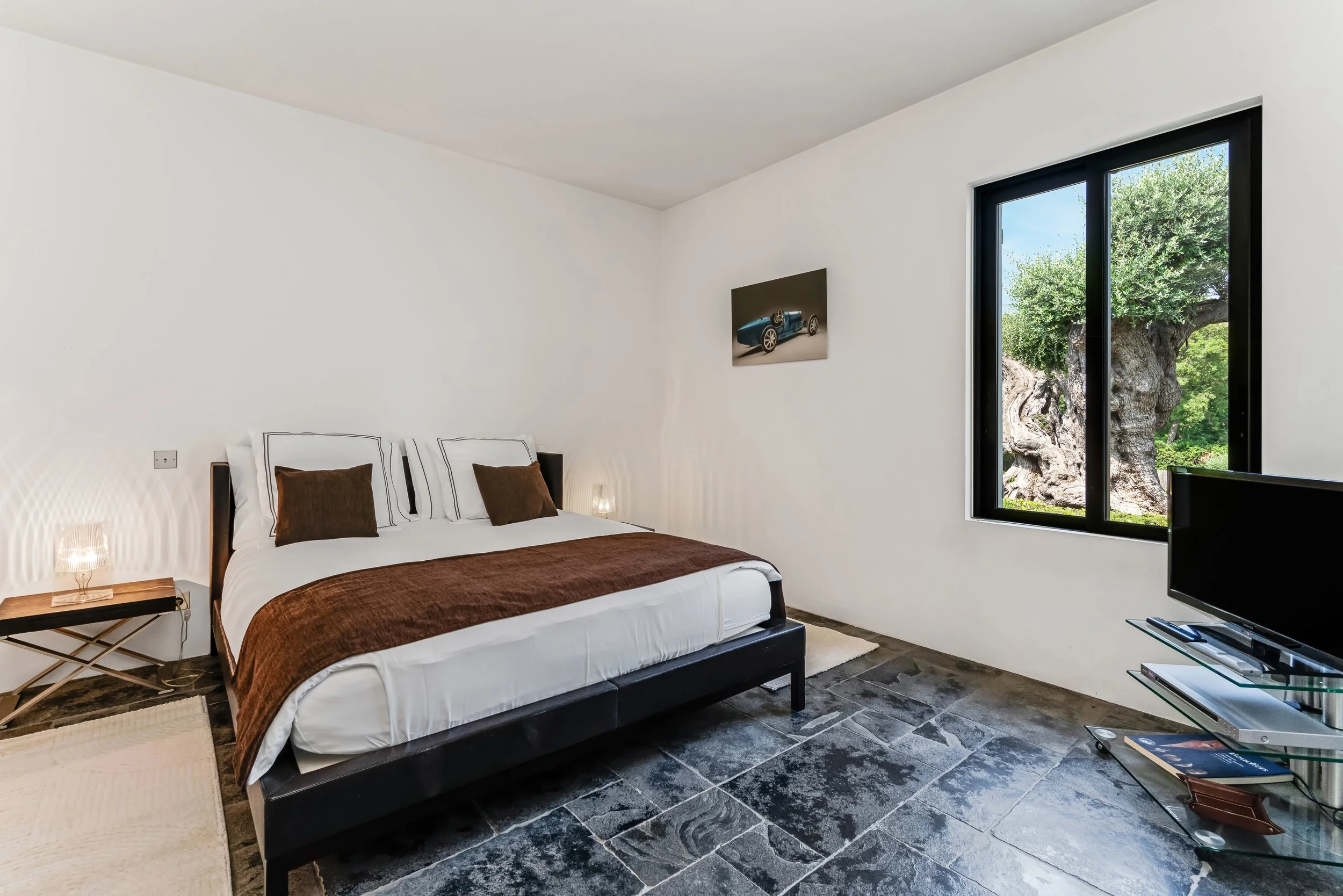 A bedroom with a large bed, brown and white bedding, a small side table with a lamp, a flat-screen TV on a glass stand, a window showing trees outside, and a wall art of a blue vintage car.