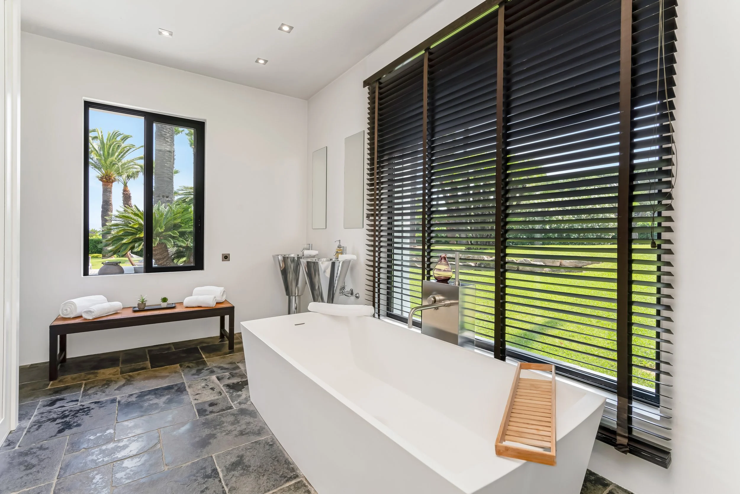Modern bathroom with a white bathtub in front of large black window blinds, a small table with towels and potted plants, a window showing palm trees outside, and natural stone flooring.