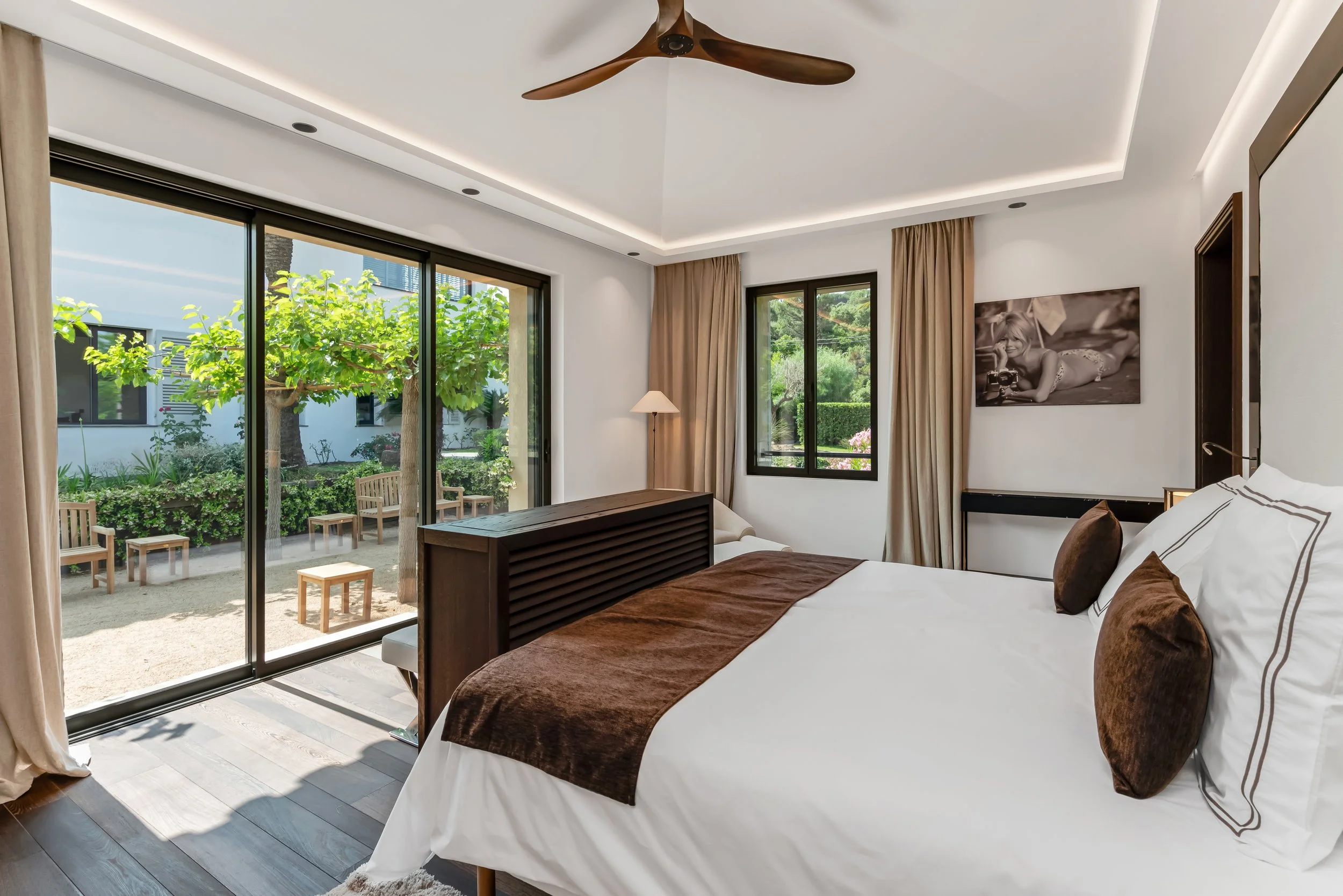 A modern bedroom with large sliding glass door opening to a garden with trees and outdoor seating, a white bed with brown pillows and a brown throw, beige curtains, a window, a black and white photograph on the wall, and a wooden ceiling fan.