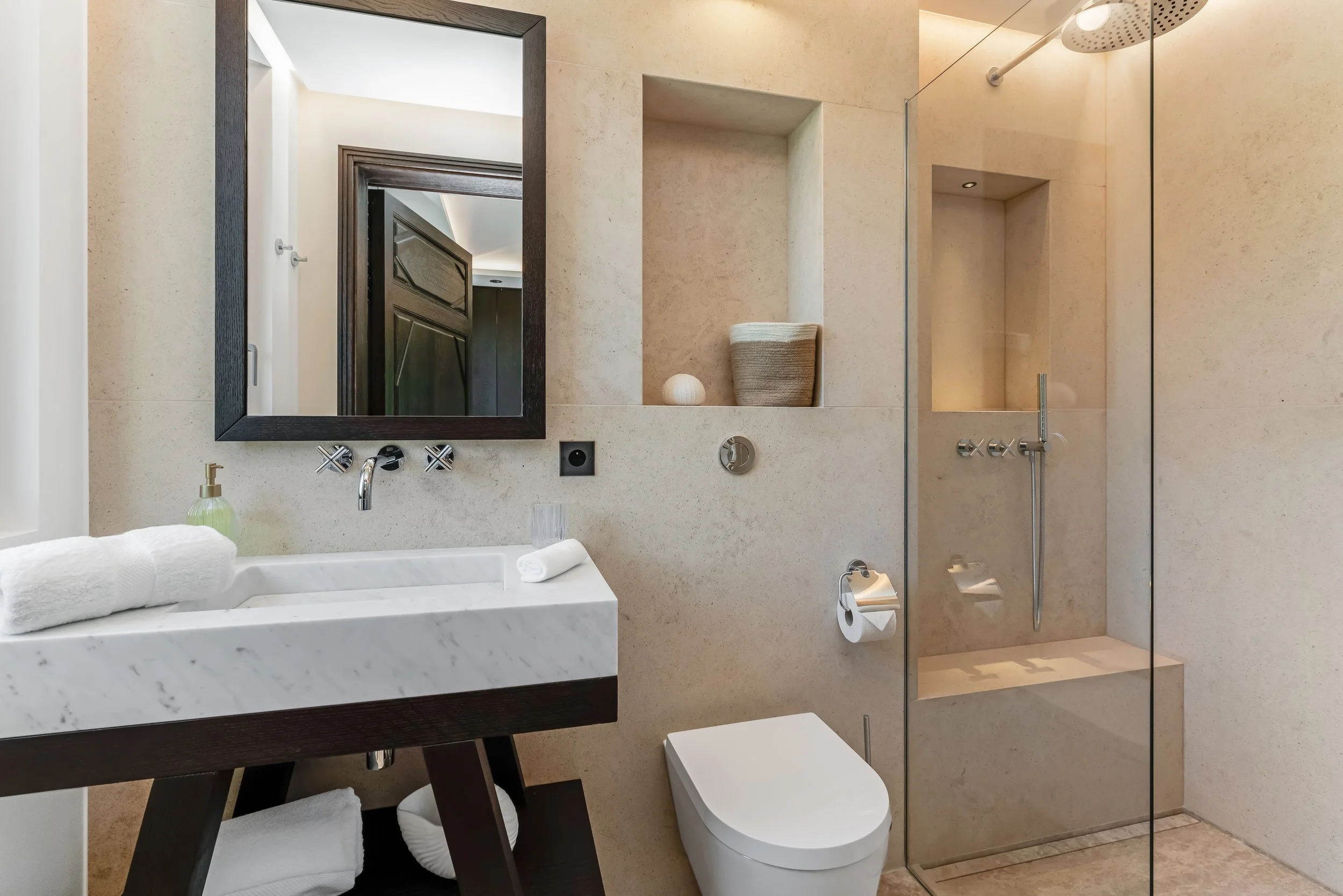 Modern bathroom with a marble sink, dark wood mirror frame, toilet, and a glass shower enclosure with a built-in shelf and rainfall showerhead.