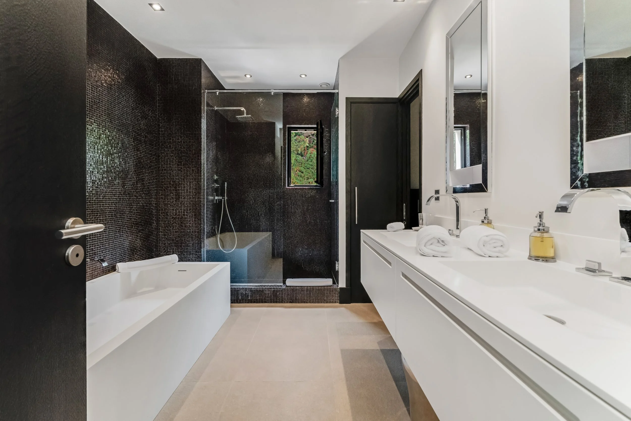 Modern bathroom with black tiled shower area, white bathtub, double white sink vanity with large mirror, towels, soap, and window with greenery outside.
