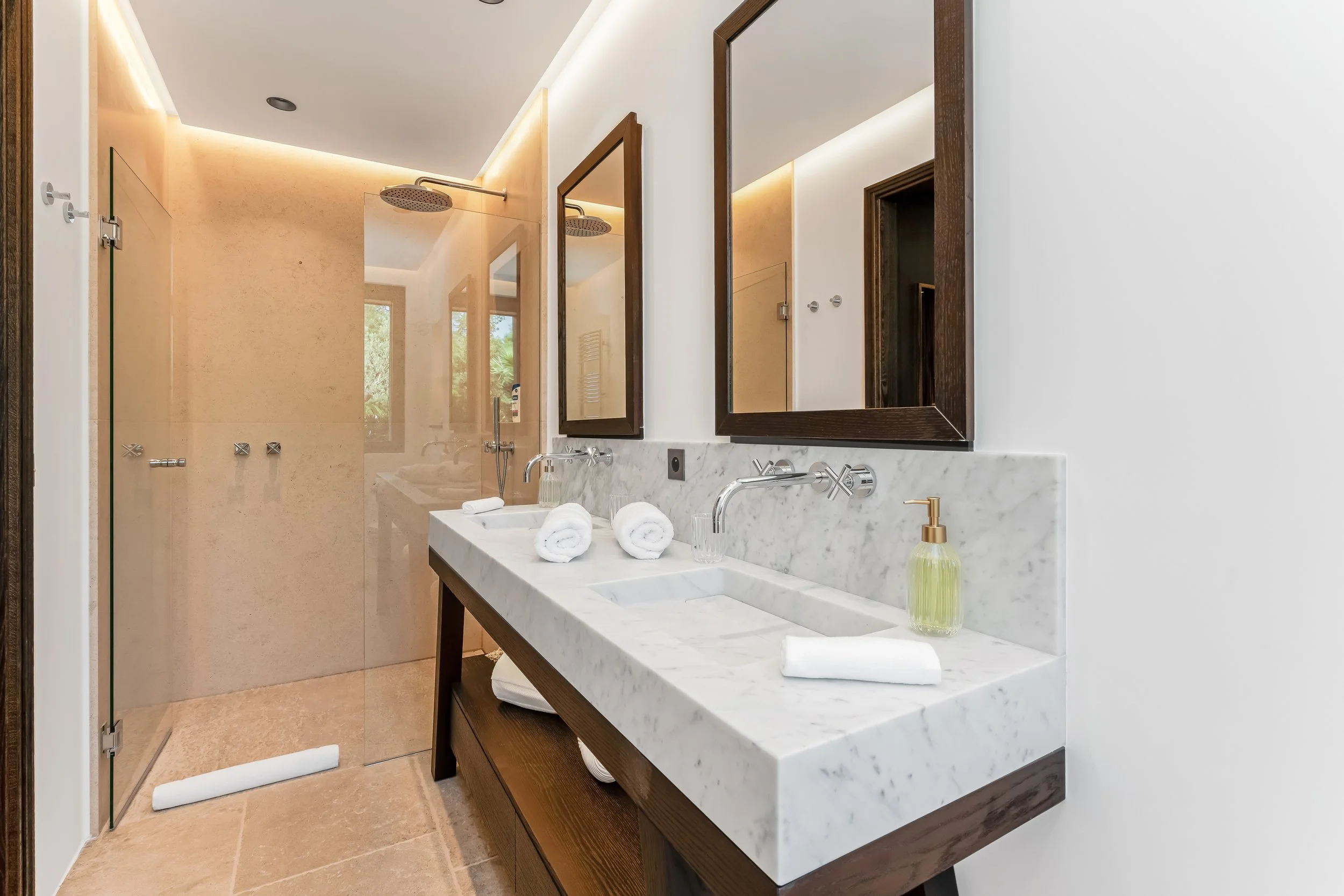 Modern bathroom with double marble sinks, wooden framed mirrors, glass shower enclosure, towels, hand soap, and natural light from windows.