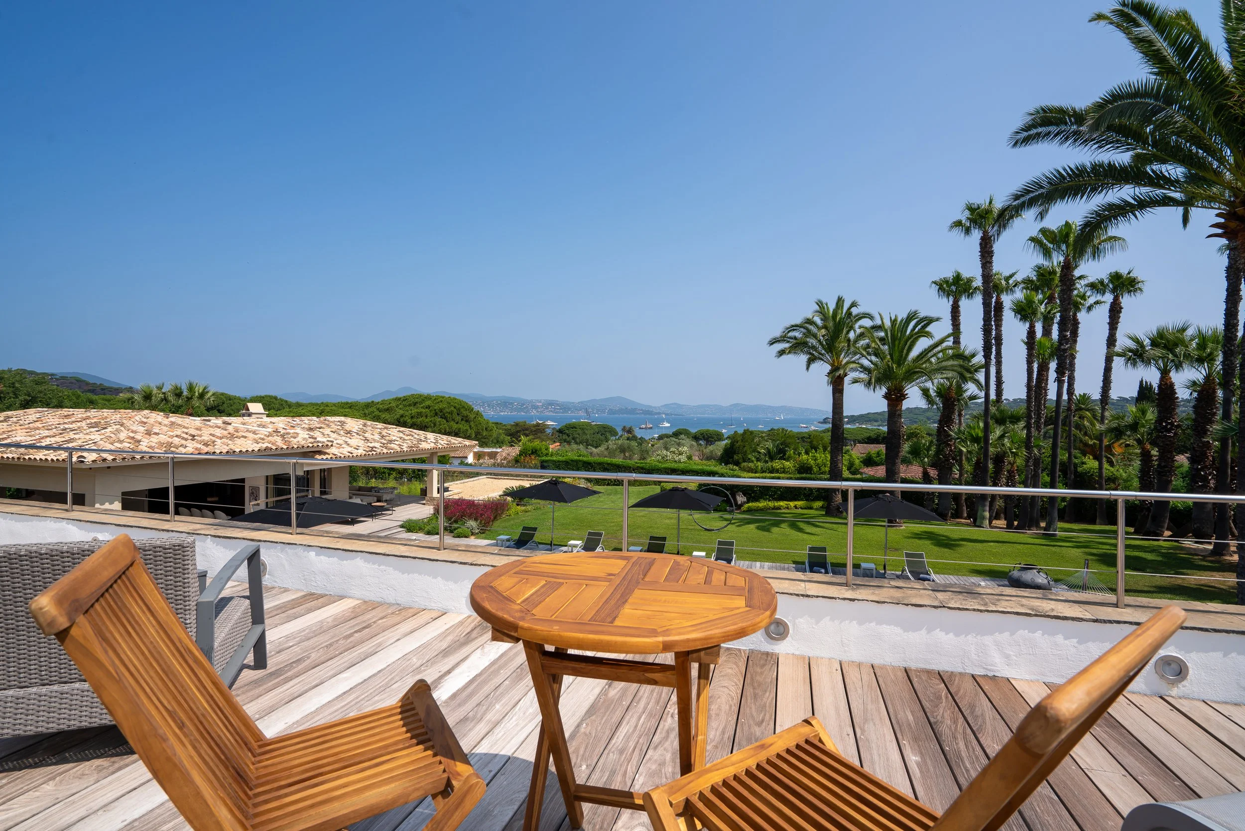 Patio with wooden chairs and table overlooking a lush green garden with palm trees, a tiled roof building, and a distant view of water and mountains under a clear blue sky.