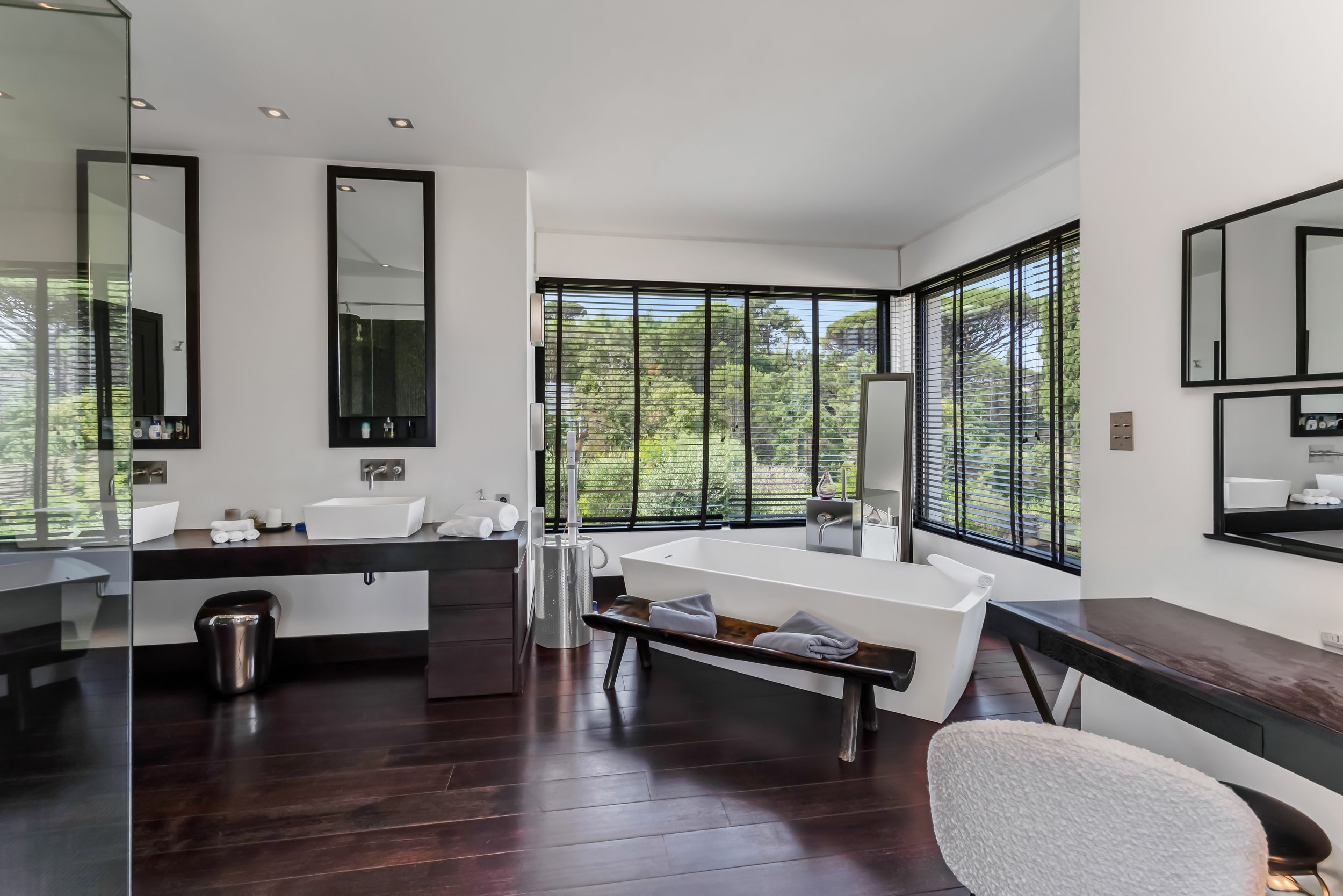 Modern bathroom with large windows, dark wood floors, white bathtub, double sink vanity, and seating area.