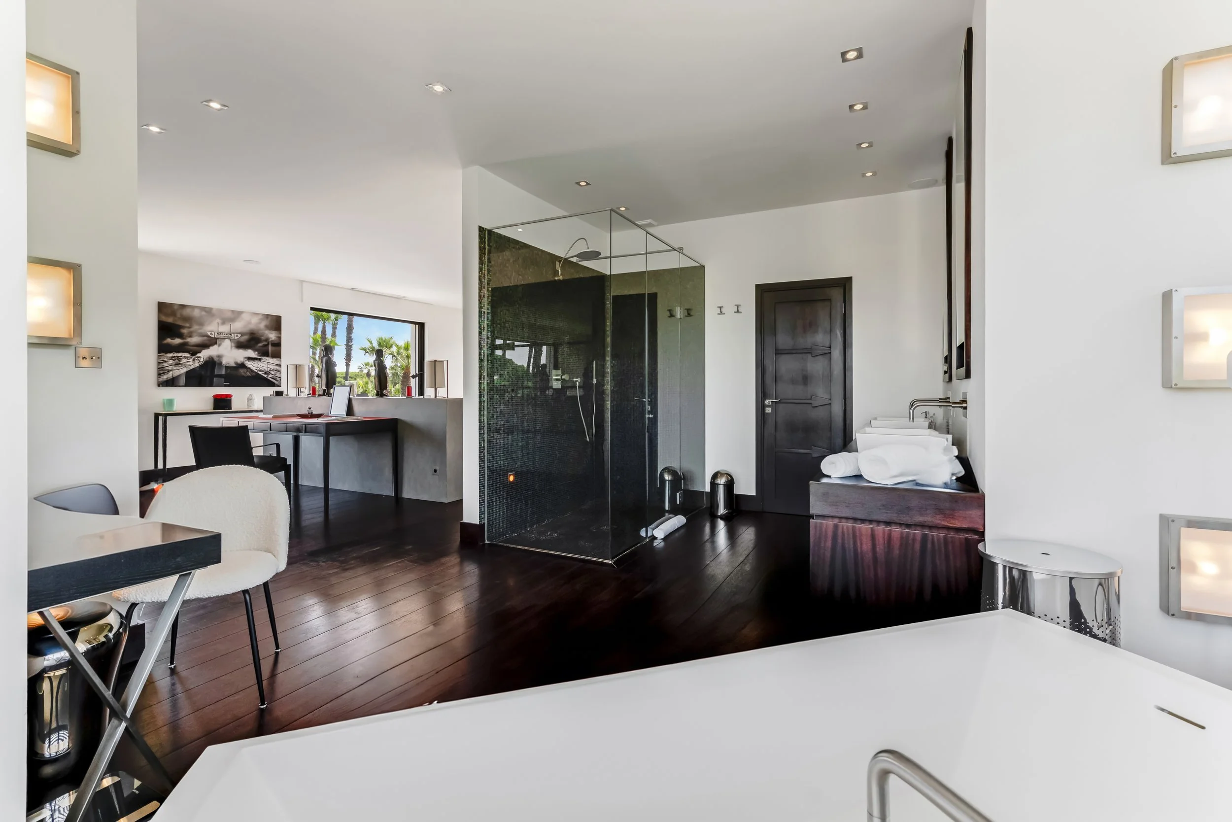 Modern bathroom and living area with dark wood flooring, a glass shower enclosure, a black door, and a white bathtub in the foreground. The living space includes a desk with a computer, a television, chairs, and windows showing palm trees outside.