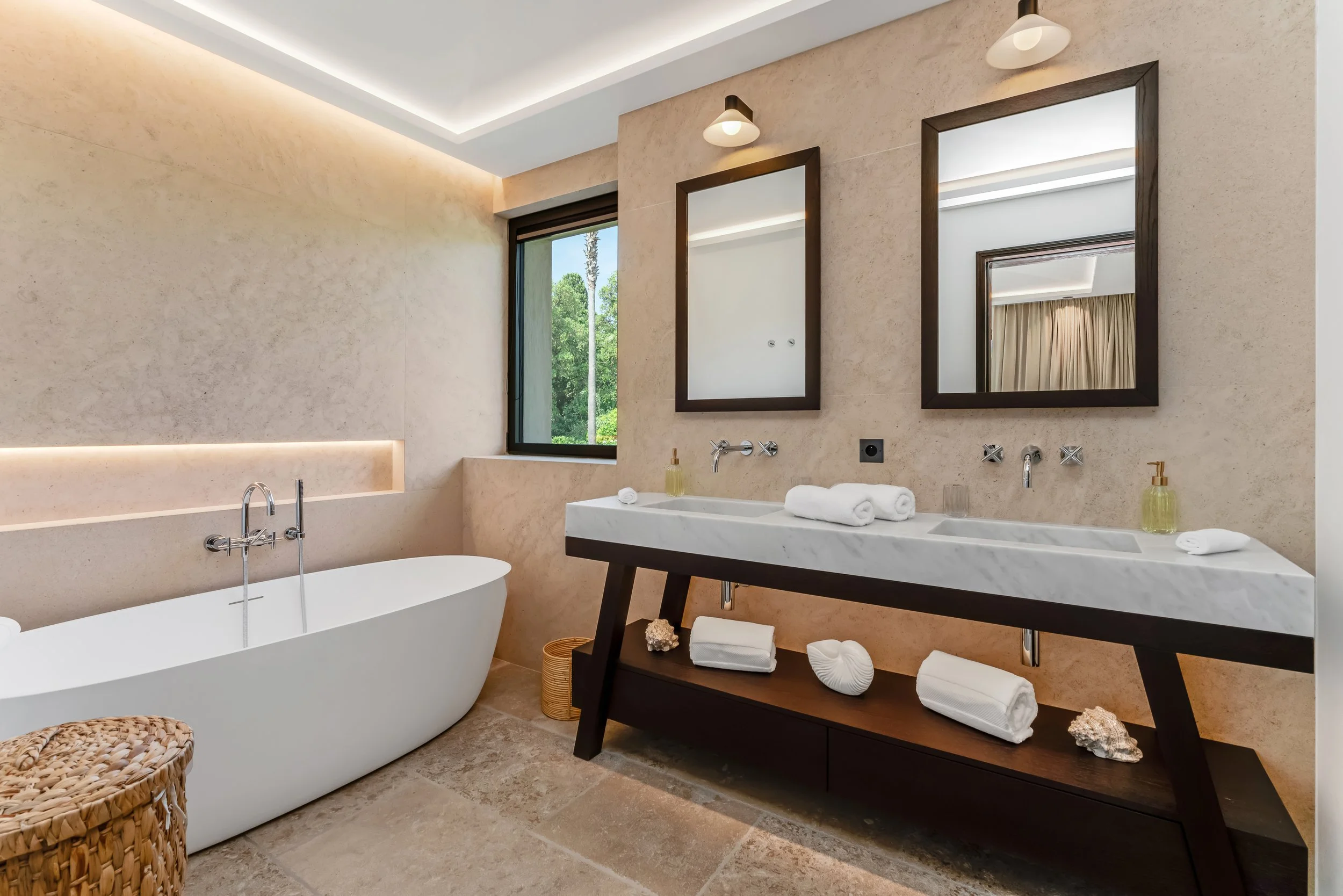 Modern bathroom with a white freestanding bathtub, double sink vanity with two mirrors, wood accents, and a window showing greenery outside