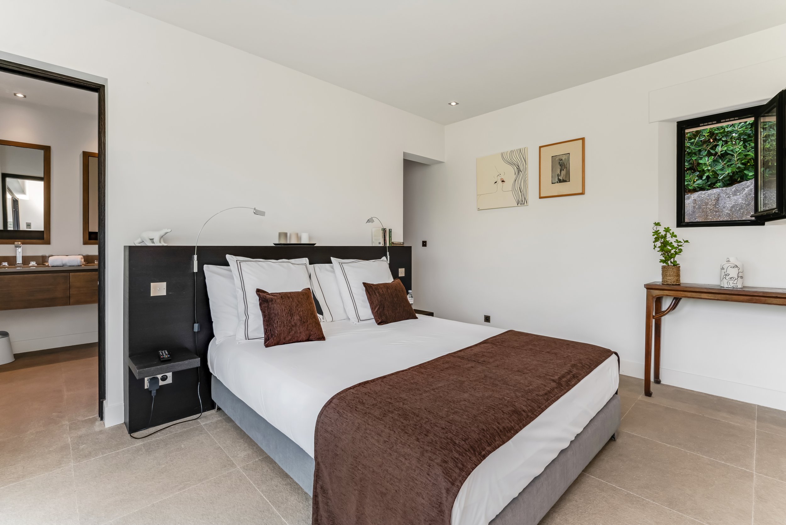 A modern hotel room with a bed having white linens, brown pillows, and a brown blanket, a black headboard, and wall-mounted reading lights. There is artwork on the white walls, a wooden table with a potted plant, and a window showing greenery outside.