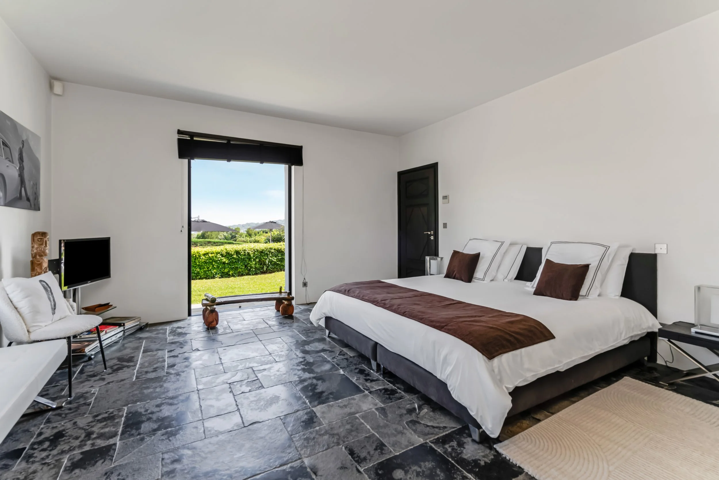 Modern hotel room with white walls, a large bed with white bedding and dark brown pillows, a small TV, a chair, a sliding glass door opening to outdoor greenery and umbrellas, and a black door.