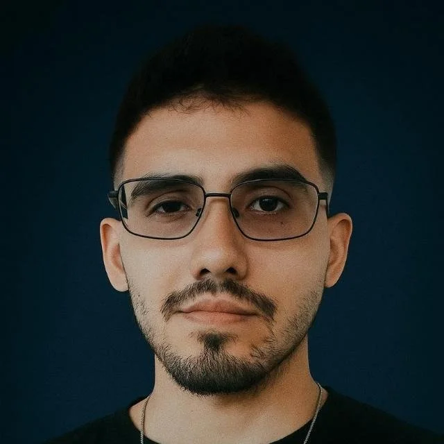 Retrato de um jovem homem de cabelo curto, barba por fazer e usando óculos quadrados com fundo azul escuro.