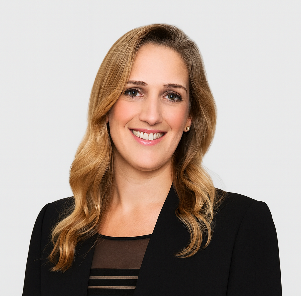 A professional woman with long blonde hair, wearing a black blazer and a sheer black top, smiling at the camera against a plain white background.