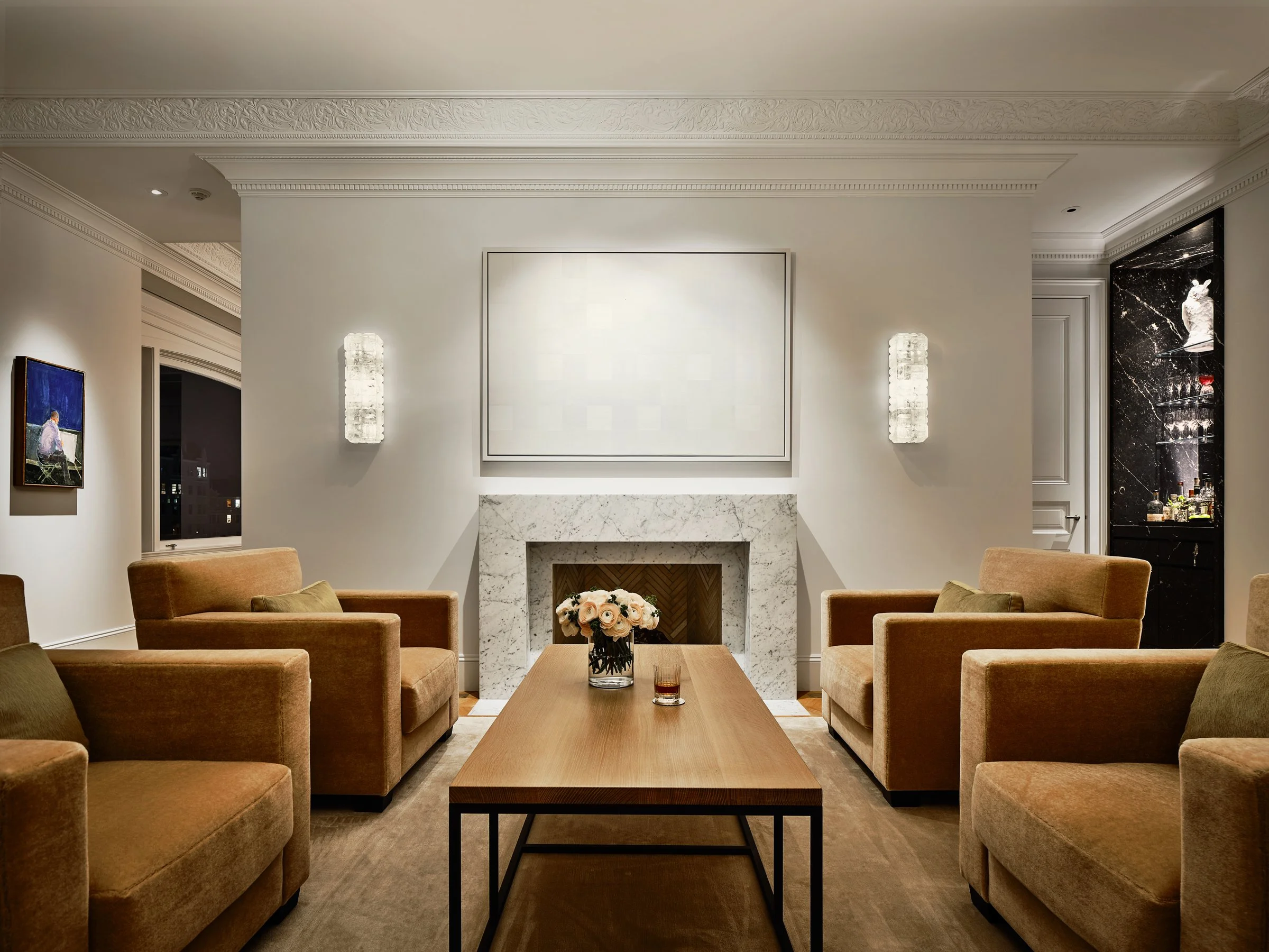 A living room furnished with six tan armchairs, a wooden coffee table with a flower vase, a fireplace with a white marble mantel, wall sconces, a blank artwork above the fireplace, and a black marble bar area on the right.