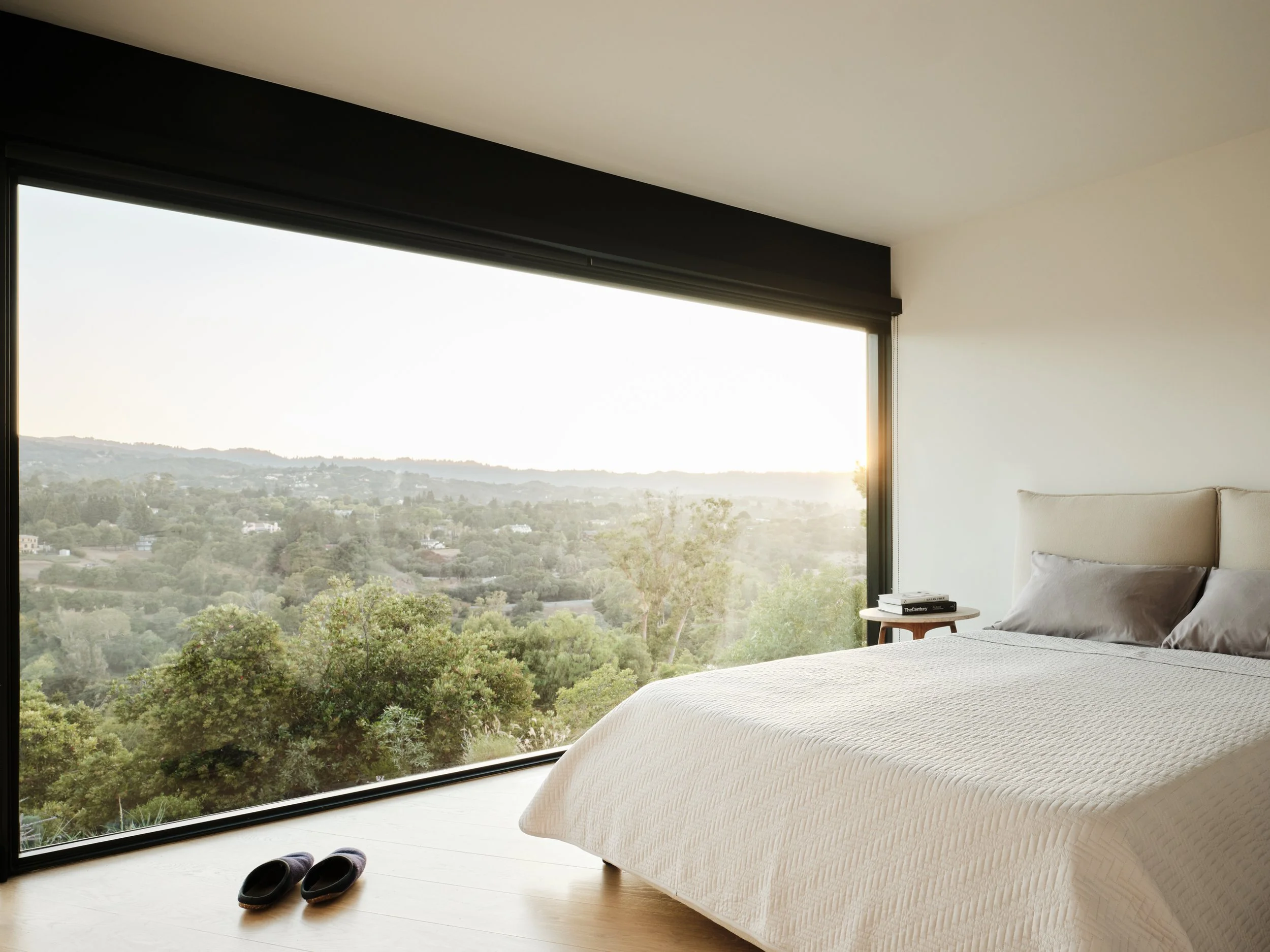 Minimalist bedroom with large window showing a scenic view of trees and hills at sunset, a bed with a beige headboard and gray pillows, a small side table with books, and slippers on the wooden floor.
