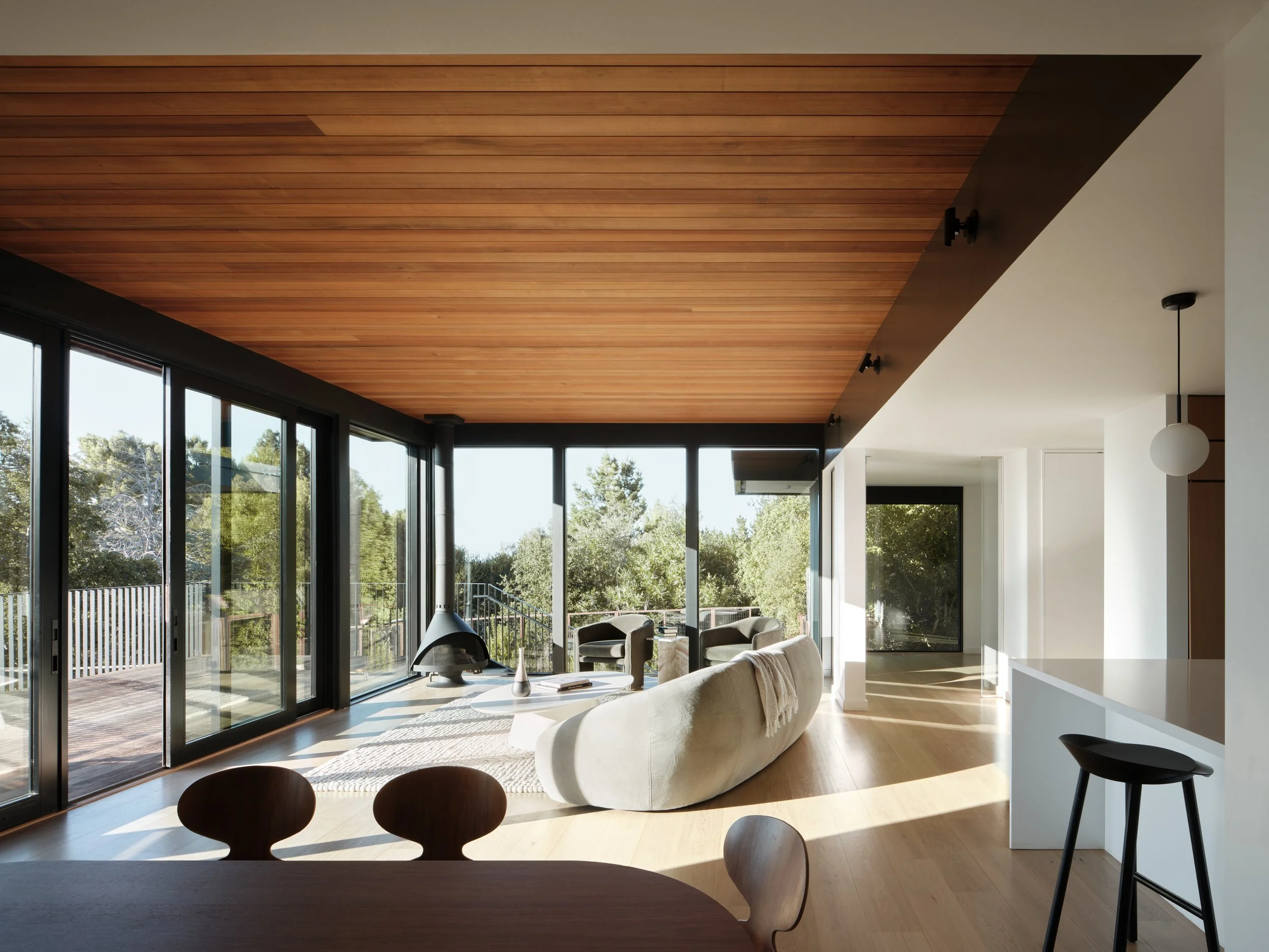 Modern living room with large glass sliding doors, wooden ceiling, and minimal furniture, looking out onto a backyard with trees.