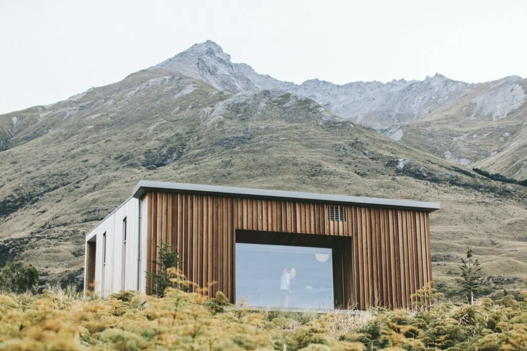 Modern wooden house with large glass window in a mountainous landscape with dry grass, small trees, and rocky mountains in the background.