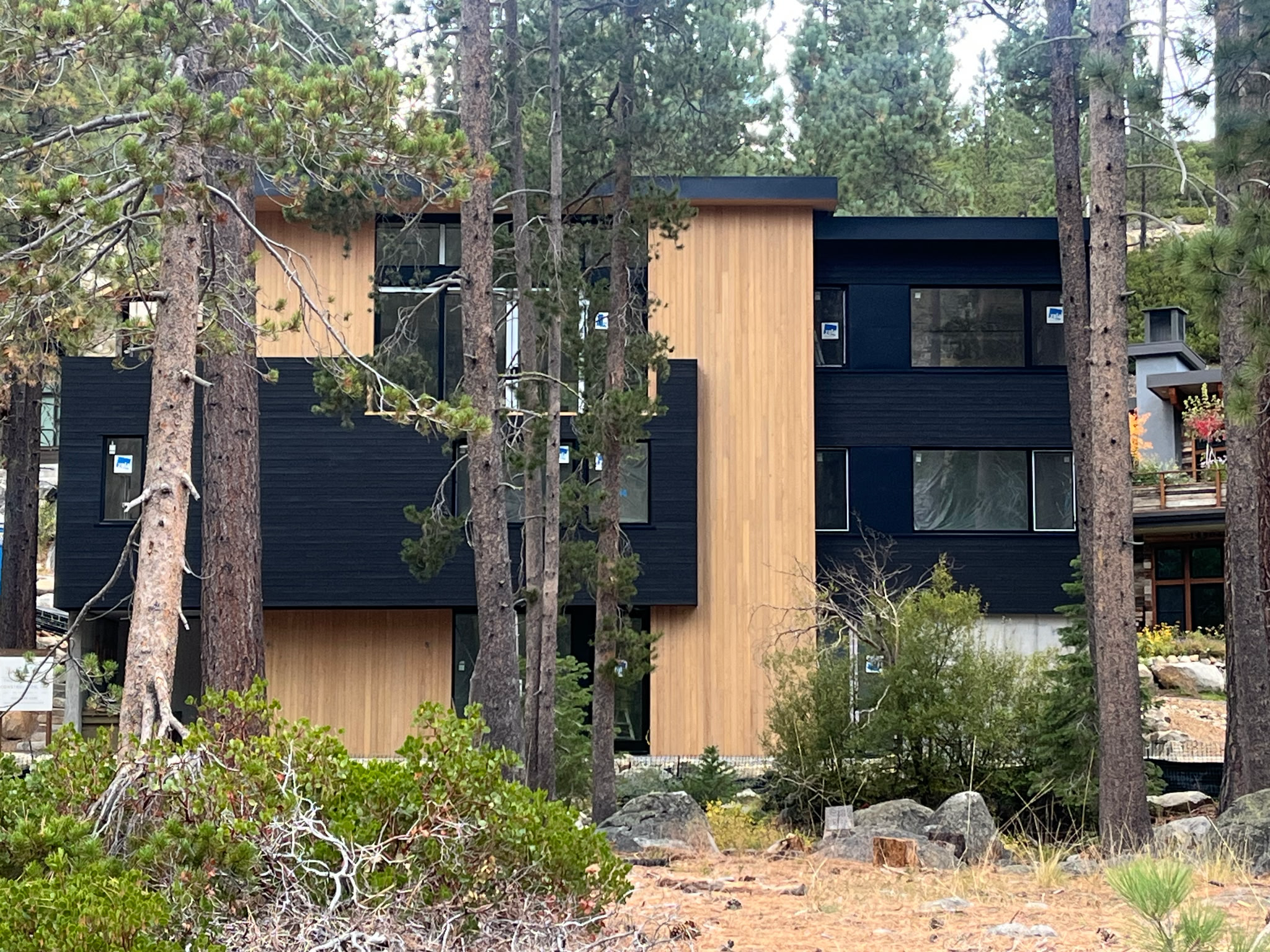 Modern multi-story house with black and wood exterior located among pine trees and rocks.