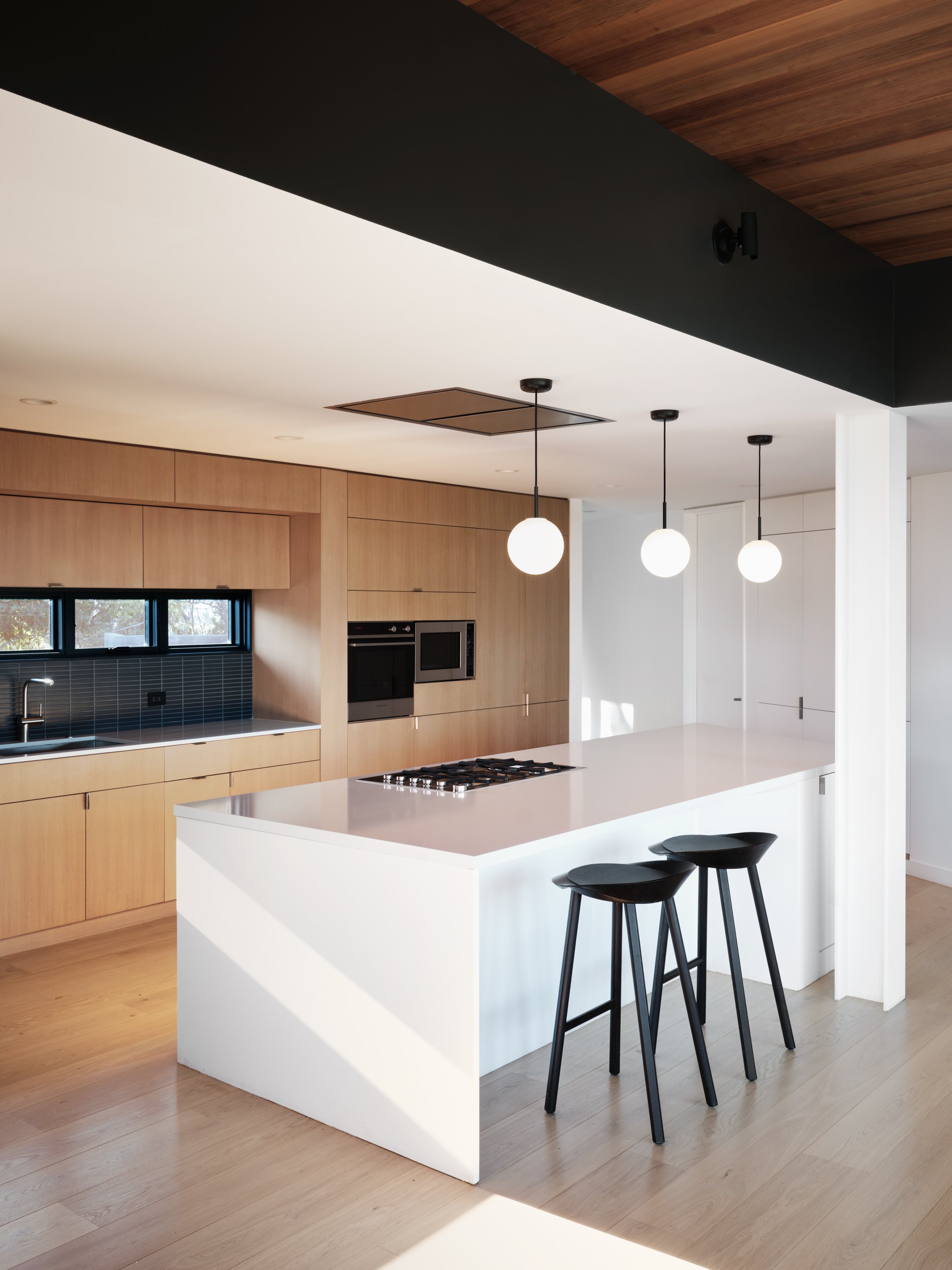 Modern kitchen with wooden cabinets, a white island with two black barstools, pendant lighting, black backsplash, and built-in appliances.