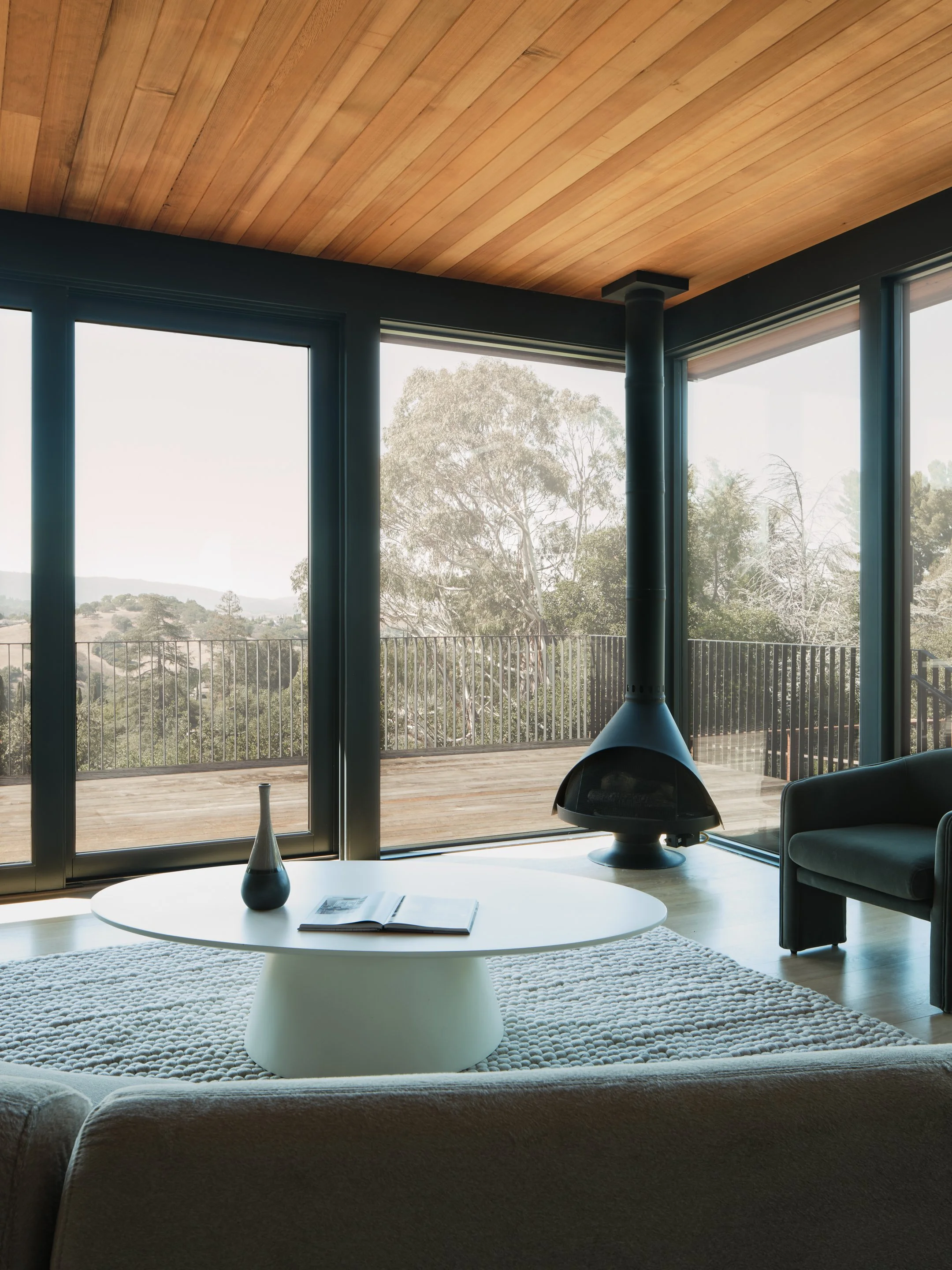 Modern living room with large glass sliding doors leading to a balcony, featuring a black hanging fireplace, white coffee table with a book and vase, and a patterned rug.