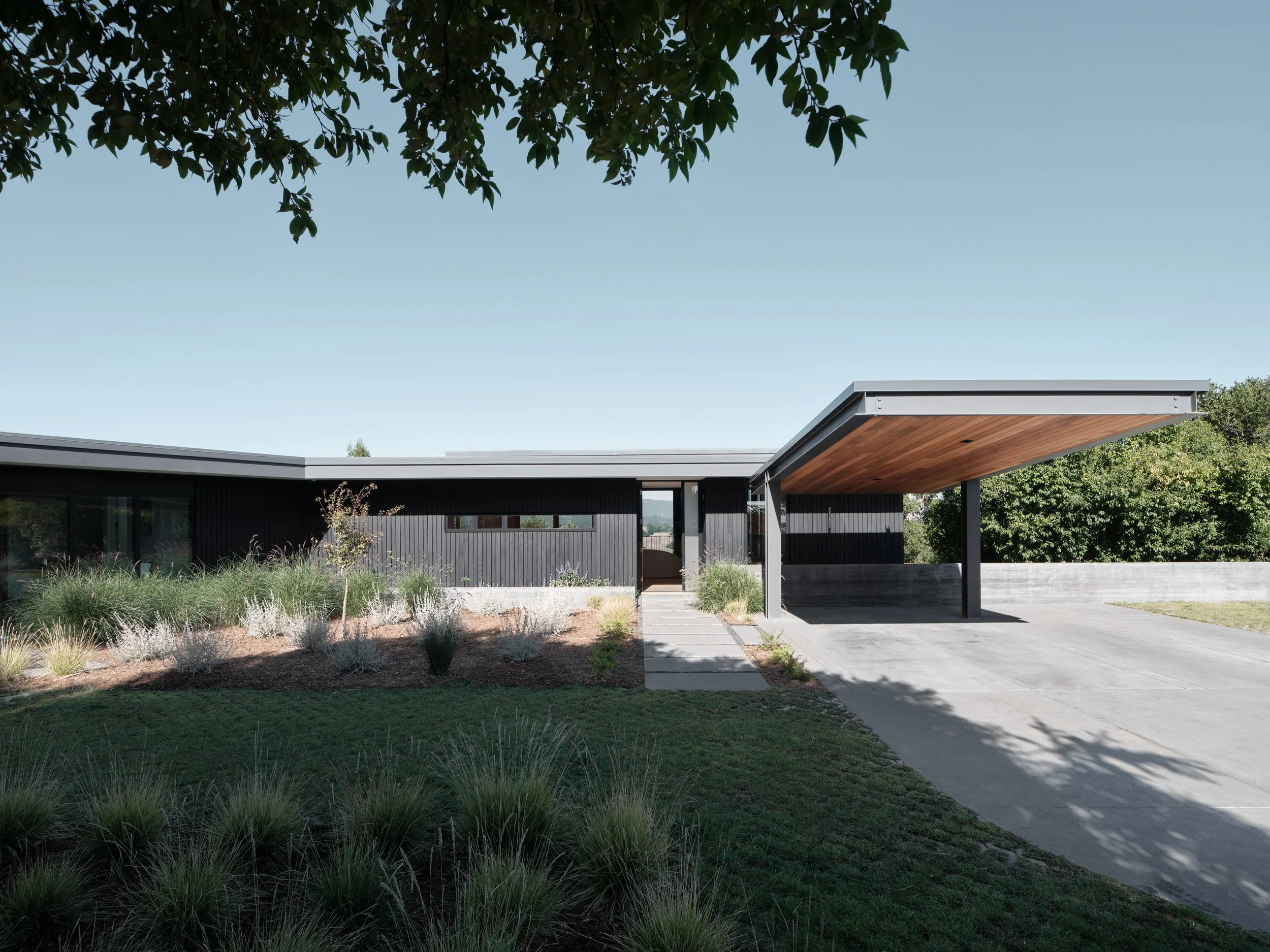 Modern single-story house with black exterior walls, large windows, and a carport with a wooden ceiling, surrounded by landscaped yard and trees.