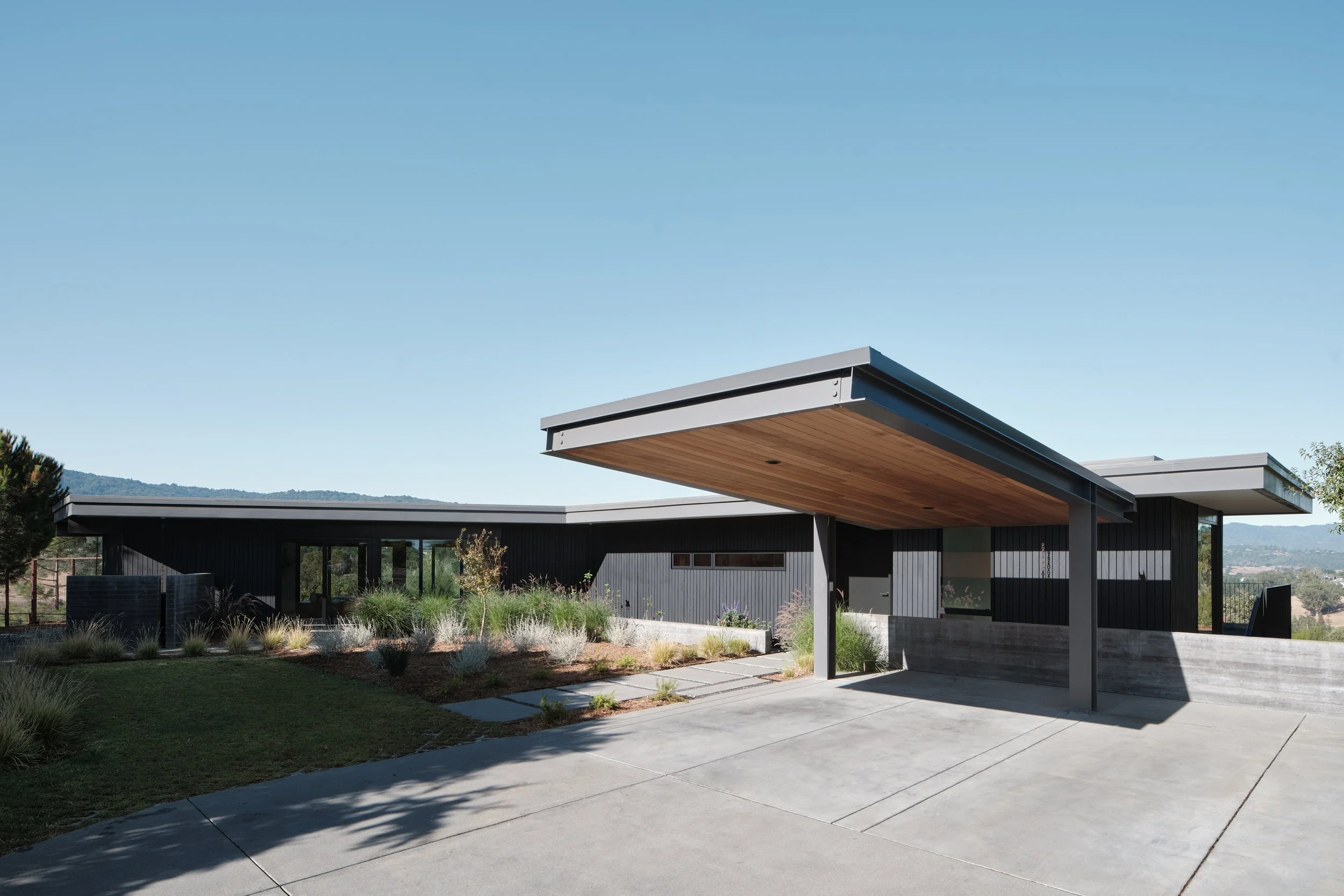 Modern single-story house with black exterior, flat roof, and a large wooden overhang at the entrance, surrounded by landscaped yard and distant hills under a clear blue sky.