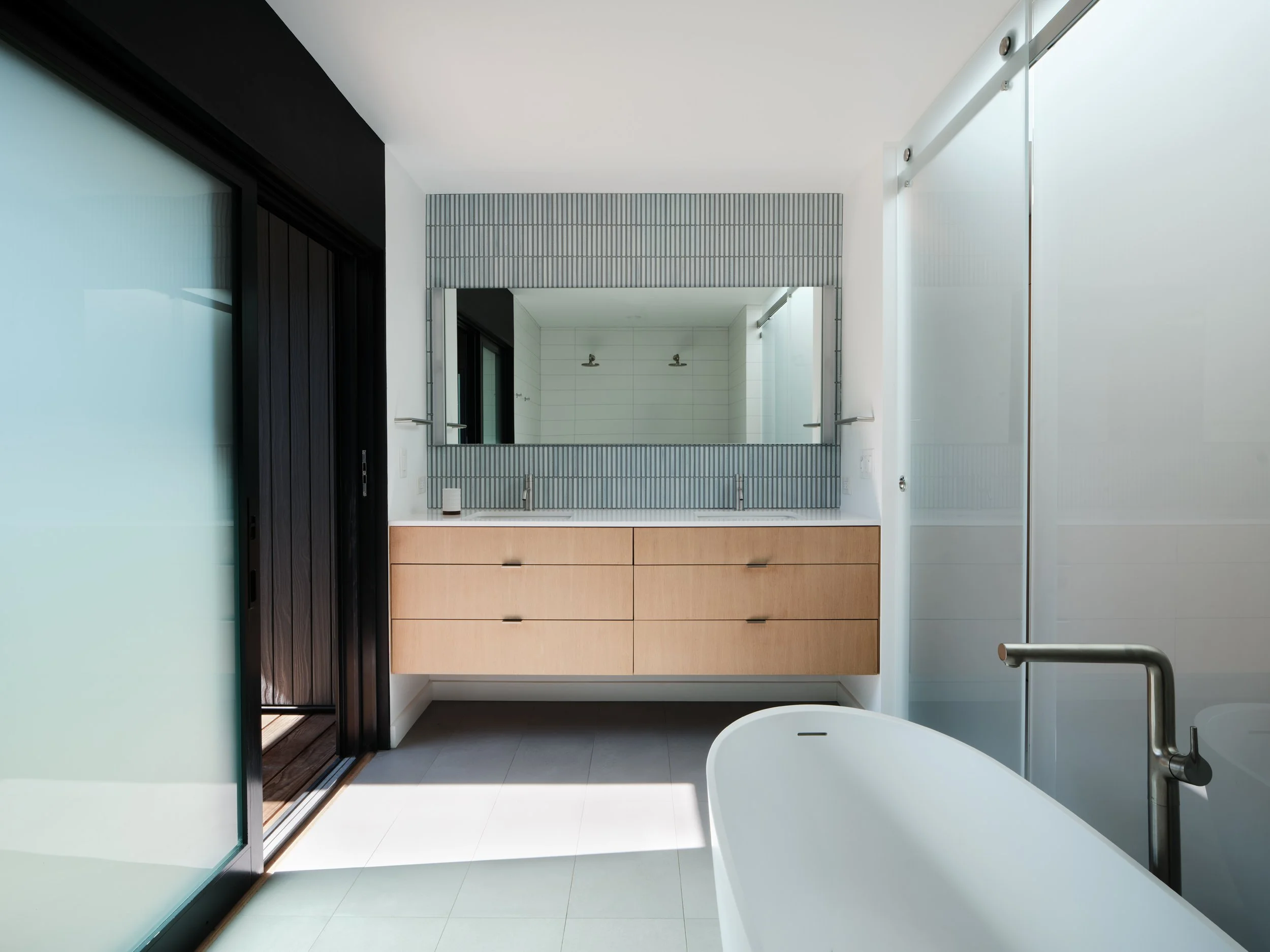 Modern bathroom with a white freestanding bathtub, a floating wooden vanity with two sinks, a large mirror, frosted glass sliding doors, and a walk-in shower with white tiles.