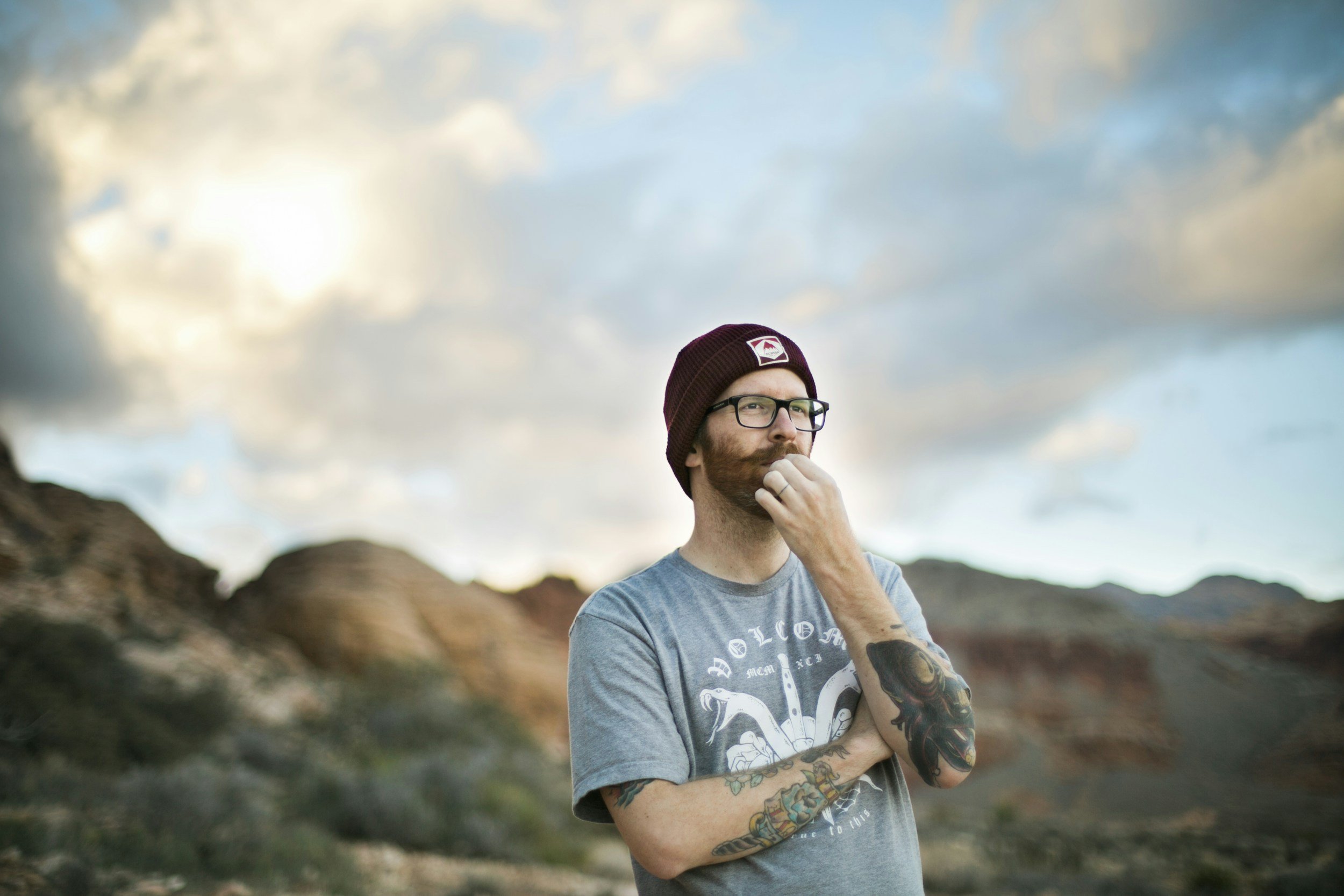 A man with tattoos and glasses wearing a maroon beanie and a gray T-shirt with a graphic, standing outdoors with rocky formations and a cloudy sky in the background, in a contemplative pose.
