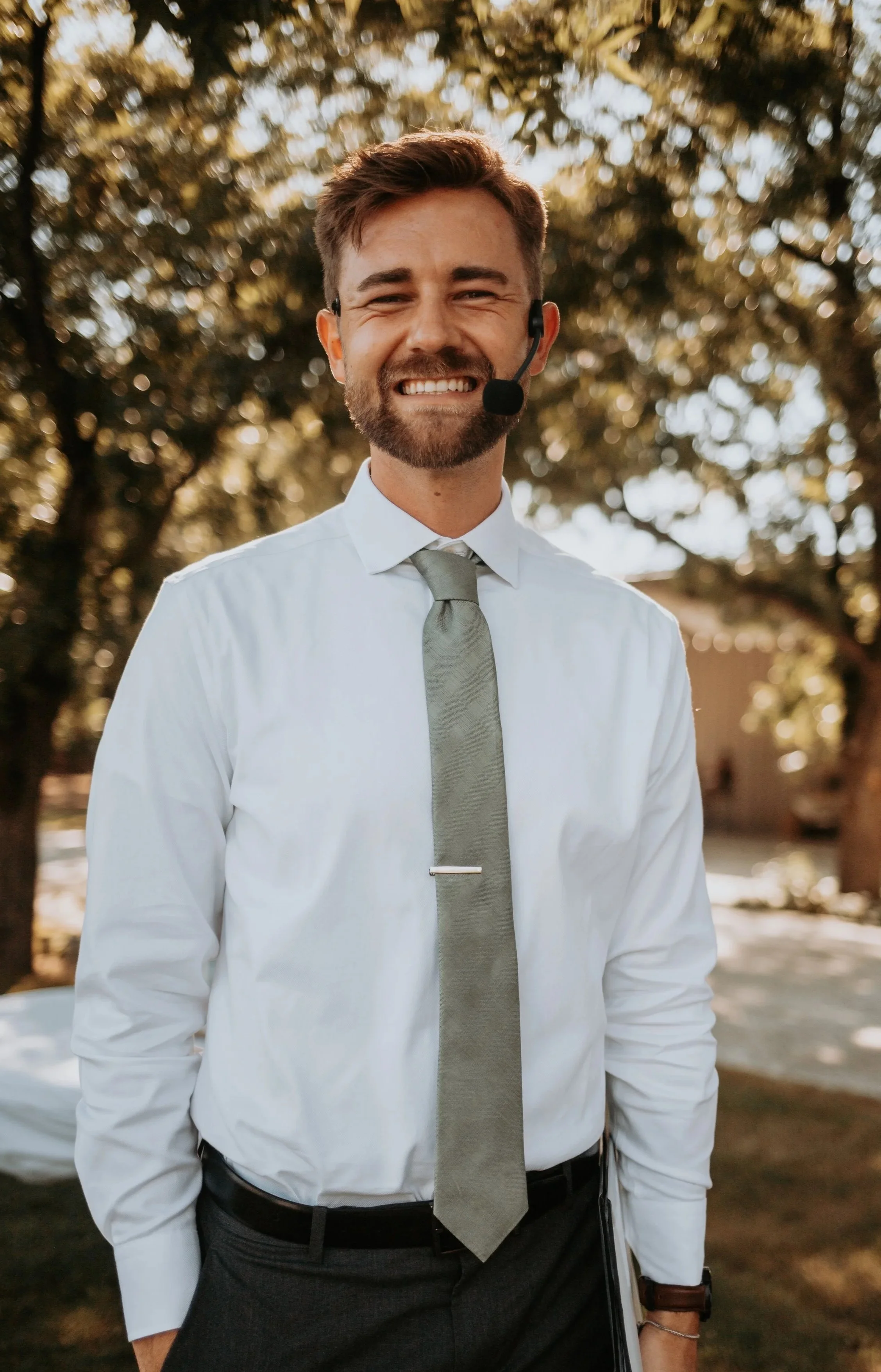 A man in formal attire wearing a white shirt, light gray tie, and a headset, standing outdoors with a background of trees and sunlight.