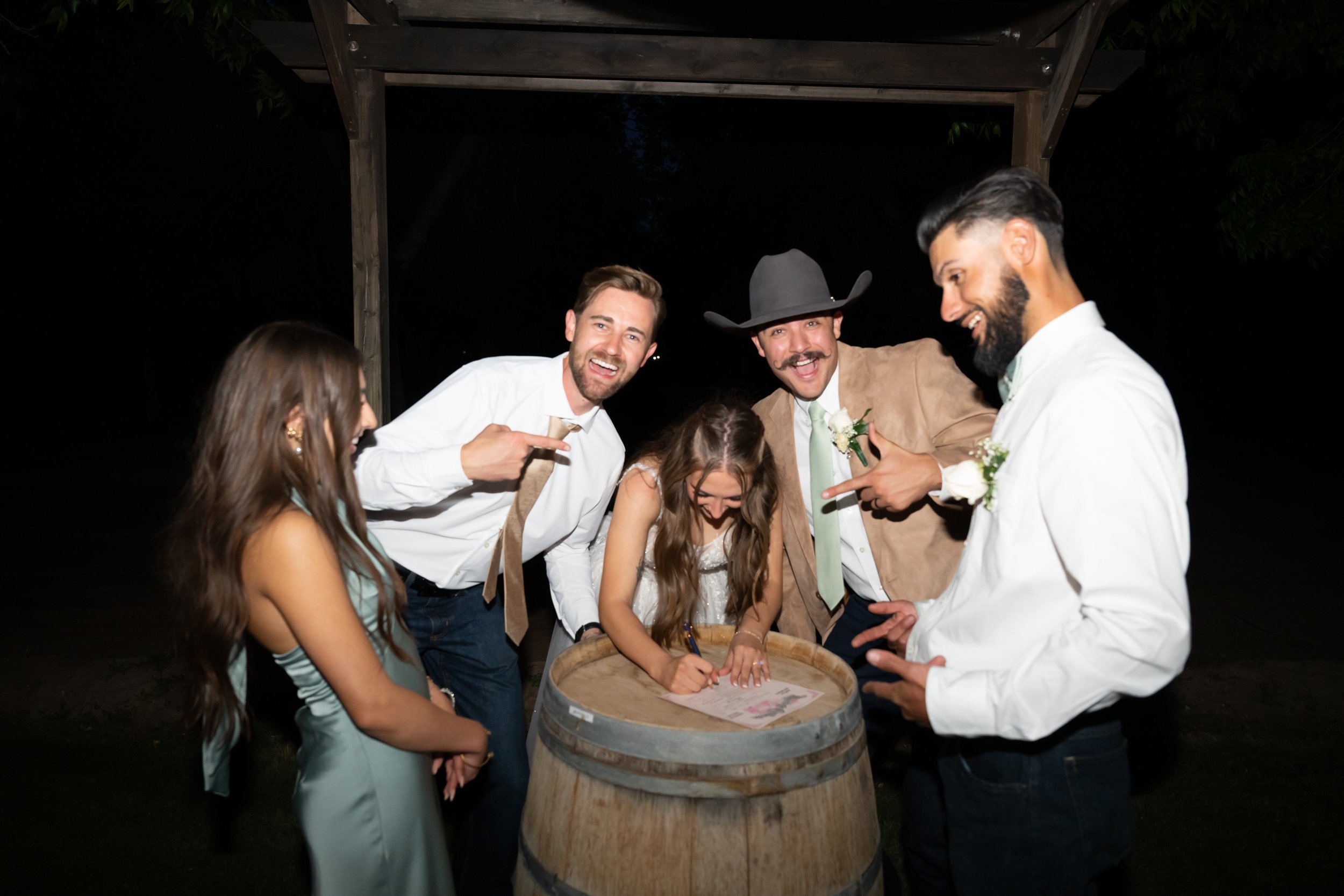 Group of five people, including a young girl, gathered around a wine barrel at night, with some signing a paper on the barrel and others pointing and smiling, outdoors at a rustic event.