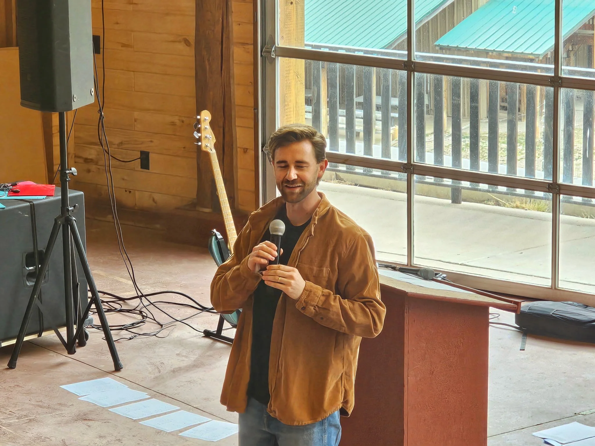 A man holding a microphone in a wooden indoor space with large front windows, a guitar leaning against the wall behind him, and audio equipment nearby.