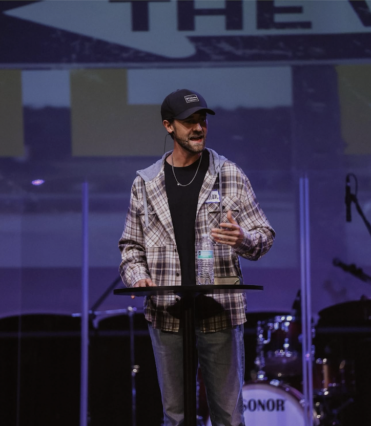 A man stands at a lectern on a stage, speaking to an audience with a microphone headset. He is wearing a black cap, a plaid jacket with a hoodie, jeans, and has a water bottle on the lectern. In the background, there is a drum set and a large screen with partially visible text.