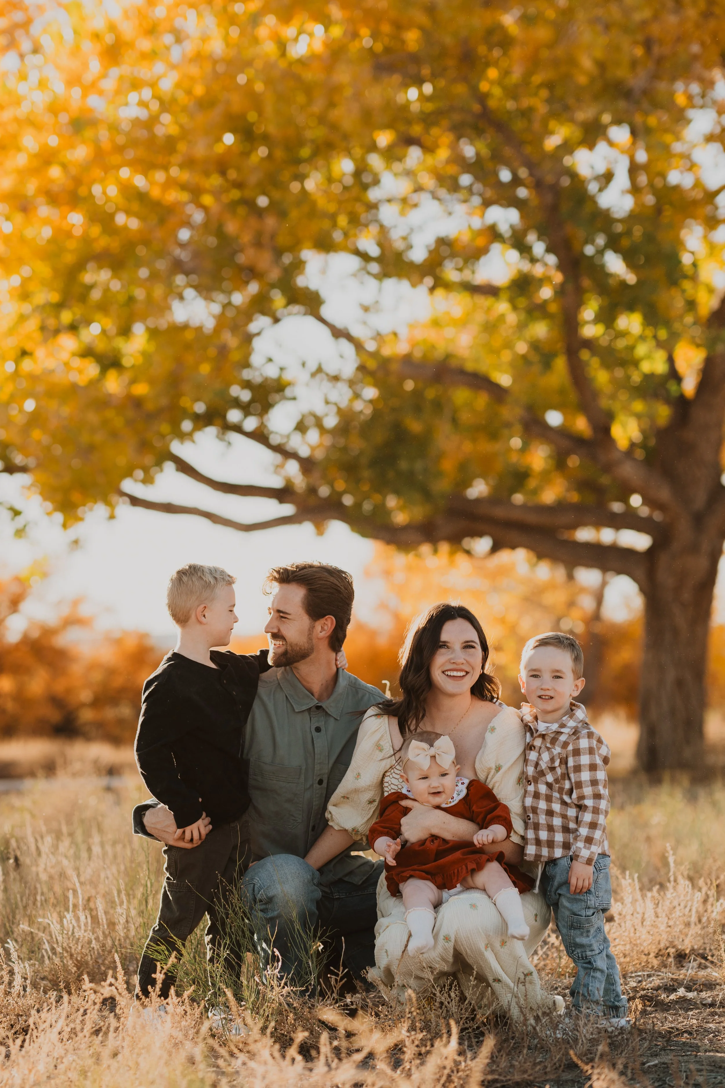 A family of five posing outdoors in a field with an autumn tree in the background, smiling and enjoying the sunlight.