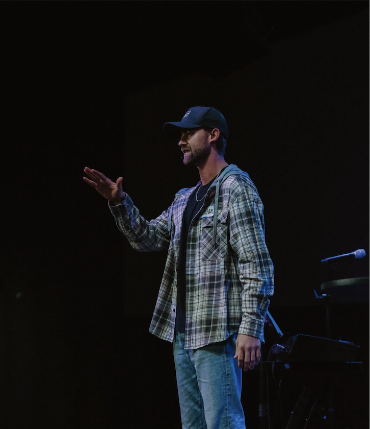 A man wearing a baseball cap, plaid shirt, and jeans gesturing with his right hand on stage with a keyboard and microphone visible.