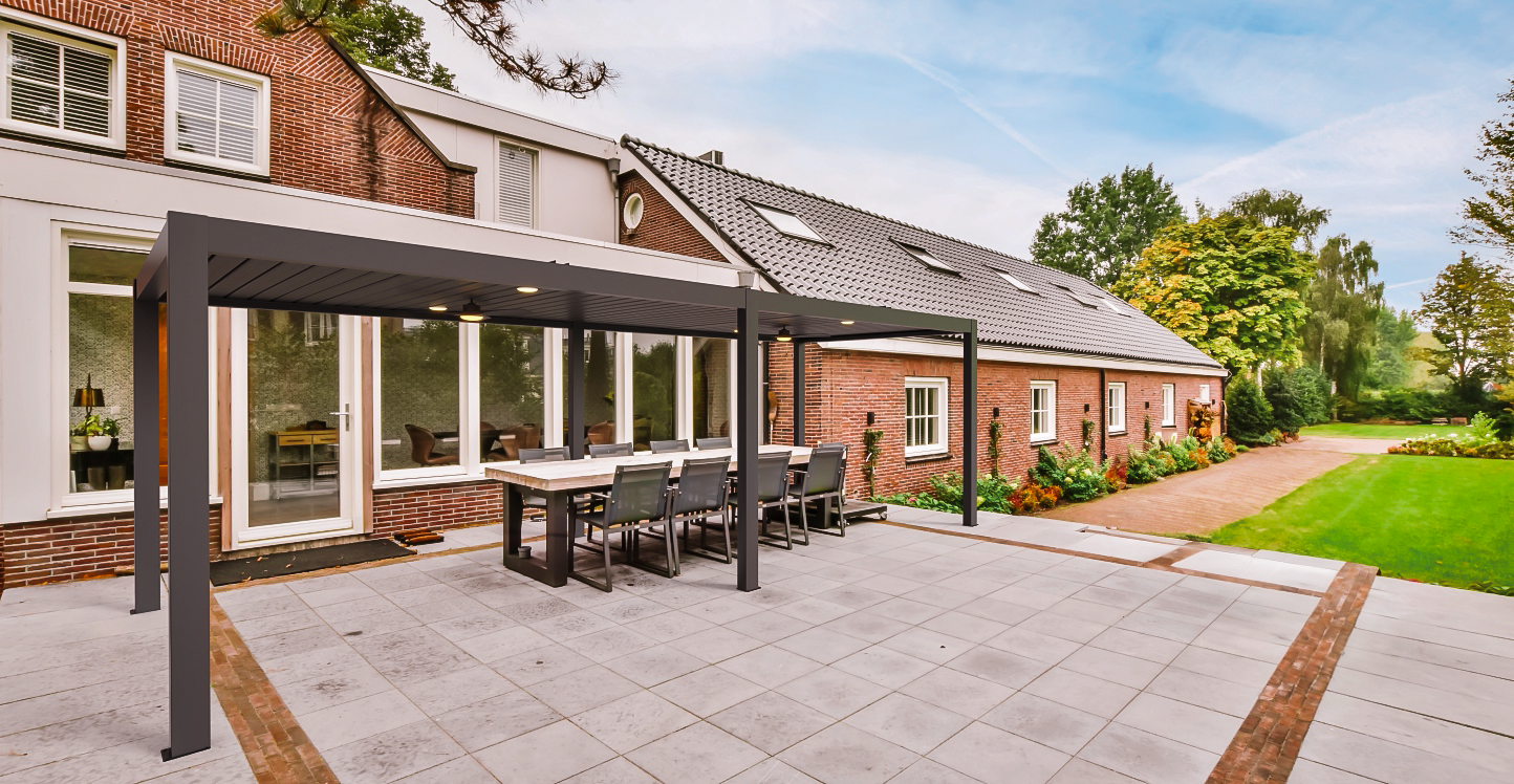 A backyard pergola attached to a brick house with a covered dining area, surrounded by a garden with green grass and trees.