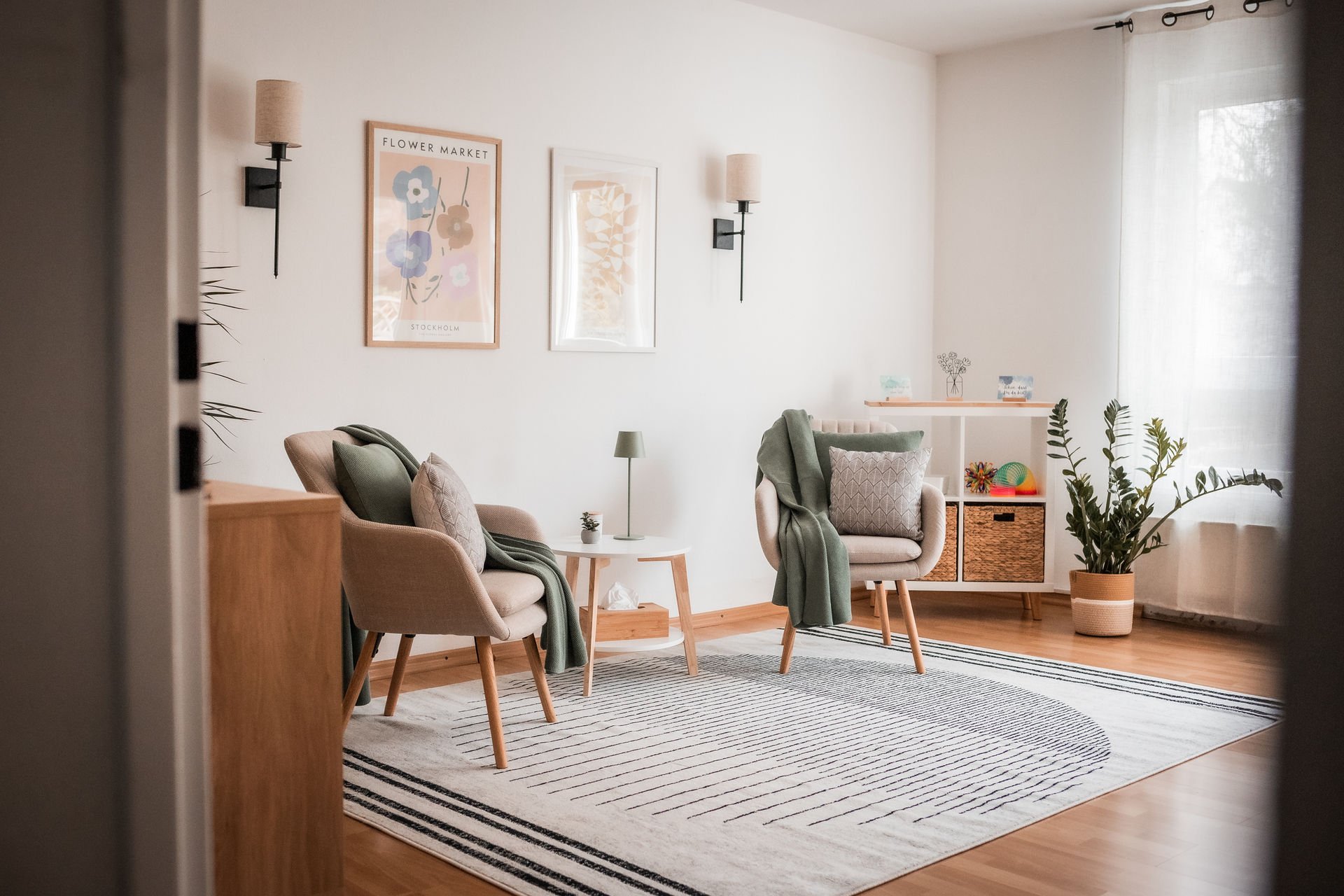 Ein hell eingerichtetes Wohnzimmer mit zwei Stühlen, einem kleinen Beistelltisch, Wandlampen, Pflanzen, Vorhängen und dekorativen Bildern an der Wand.
