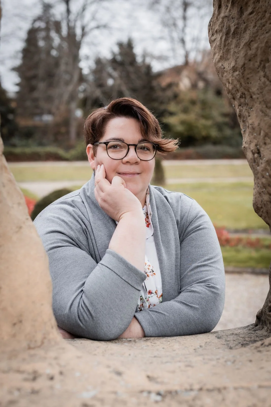 Portrai von Dr. med. Kathrin Scheffler, lächelnd, schaut durch eine Steinöfnung im Park mit Bäumen und grünem Rasen im Hintergrund. Einladen warmes Lächeln