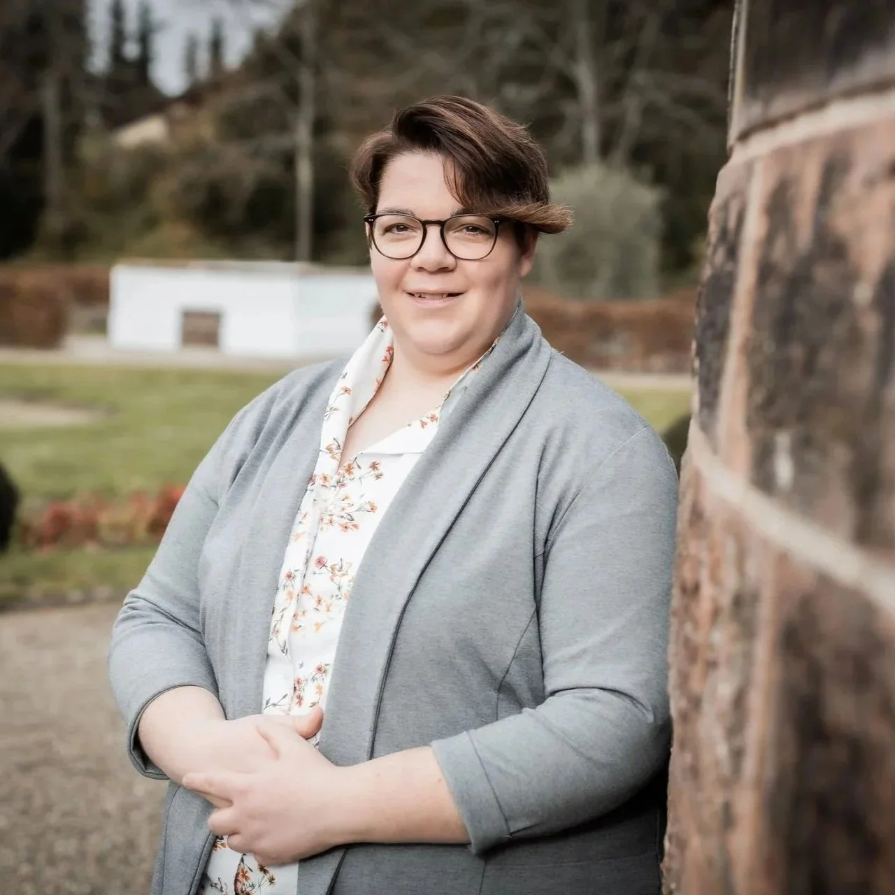 Dr. Kathrin Scheffler lehnt entspannt an einem Baumstamm im Außenbereich – symbolische Darstellung von Erdung, Ressourcenorientierung und Nervensystemregulation im Coaching.