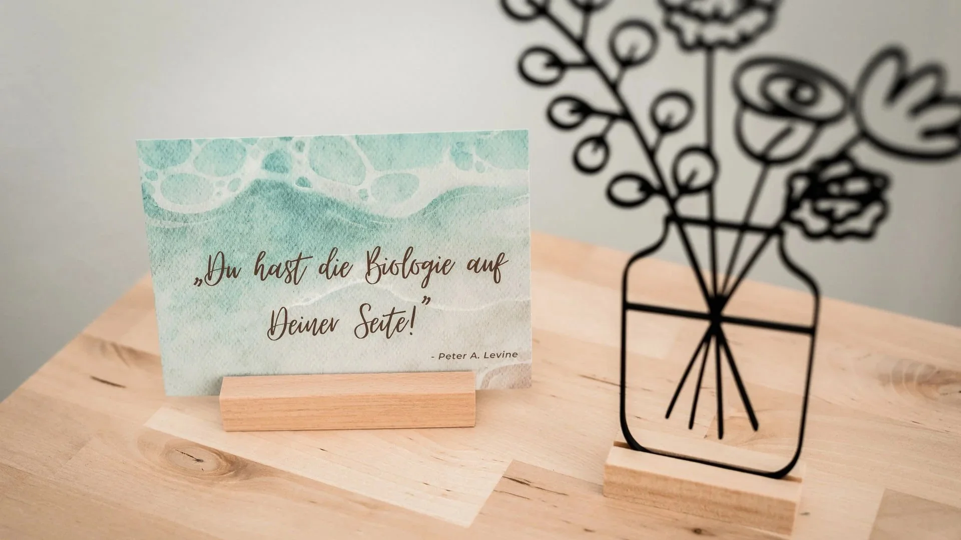 Eine stilvolle Tafel mit einem Zitat auf einer Grußkarte, daneben ein Metall-Dekorationsstück, beides auf einem Holztisch.