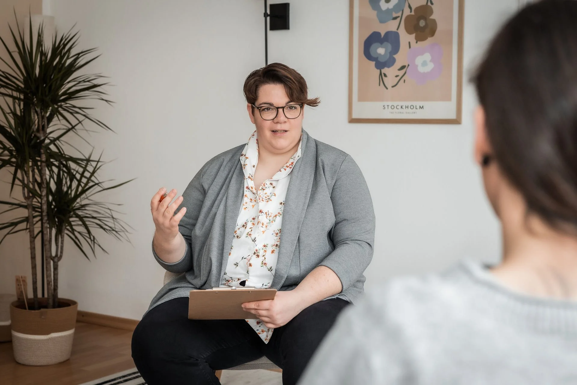 Eine Frau im Gespräch bei einer Beratungssitzung, hält einen Notizblock in der Hand, im Hintergrund ein Pflanzen und eine Wand mit einem blumenförmigen Poster mit der Aufschrift ‚Stockholm‘.