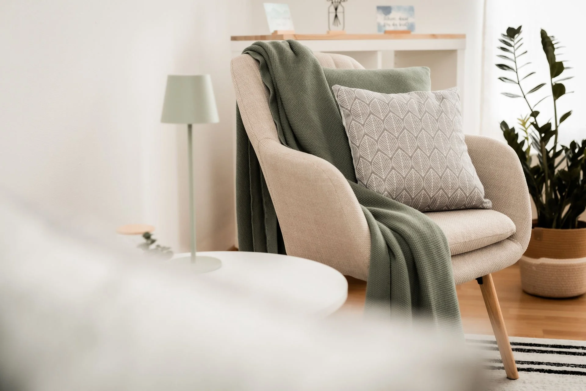 Ein gemütlicher Sessel mit Kissen und einem grünen Mantel, im Hintergrund eine Stehlampe und eine Pflanze in einem Topf in einem hellen Raum.