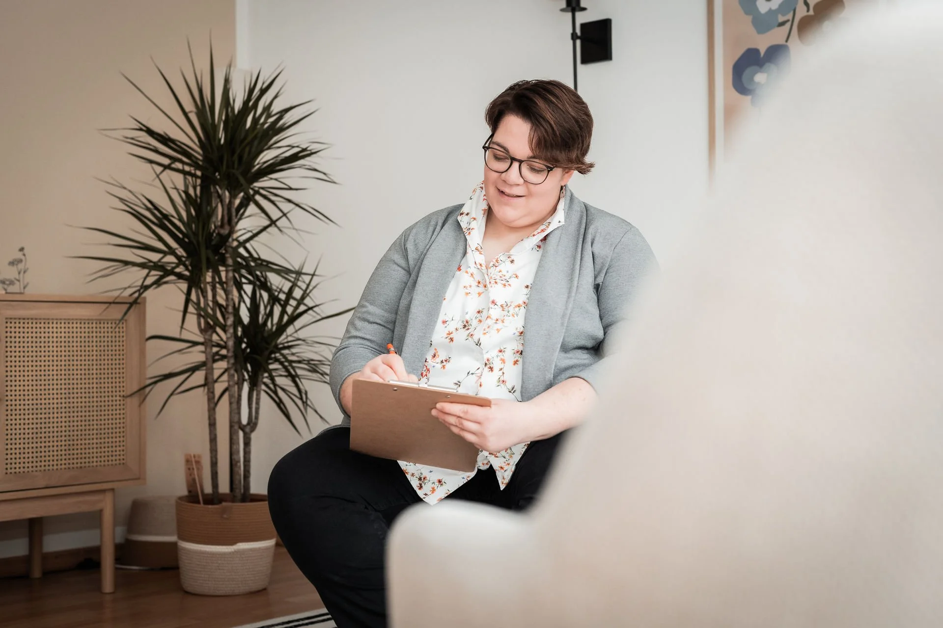 Dr. med. Kathrin Scheffler sitzt in einem Raum auf einem Stuhl, schreibt auf einem Schreibbrett, trägt eine Brille und hat kurze braune Haare, im Hintergrund sind Pflanzen und Wanddekorationen sichtbar.