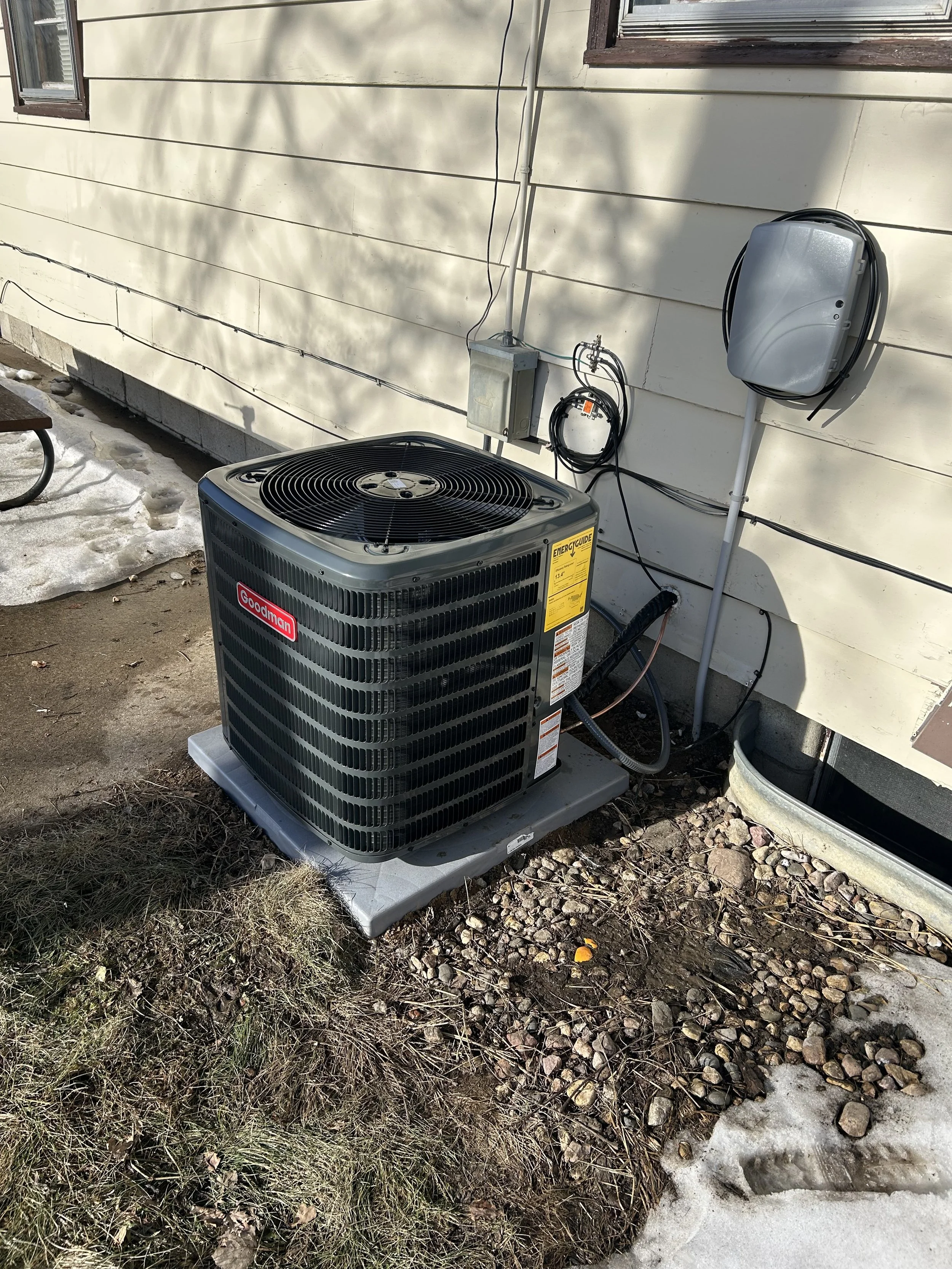 An outdoor view of a Goodman central air conditioning unit installed beside a house, with electrical connections and a grey electrical box on the wall, surrounded by patches of snow, dirt, and grass.