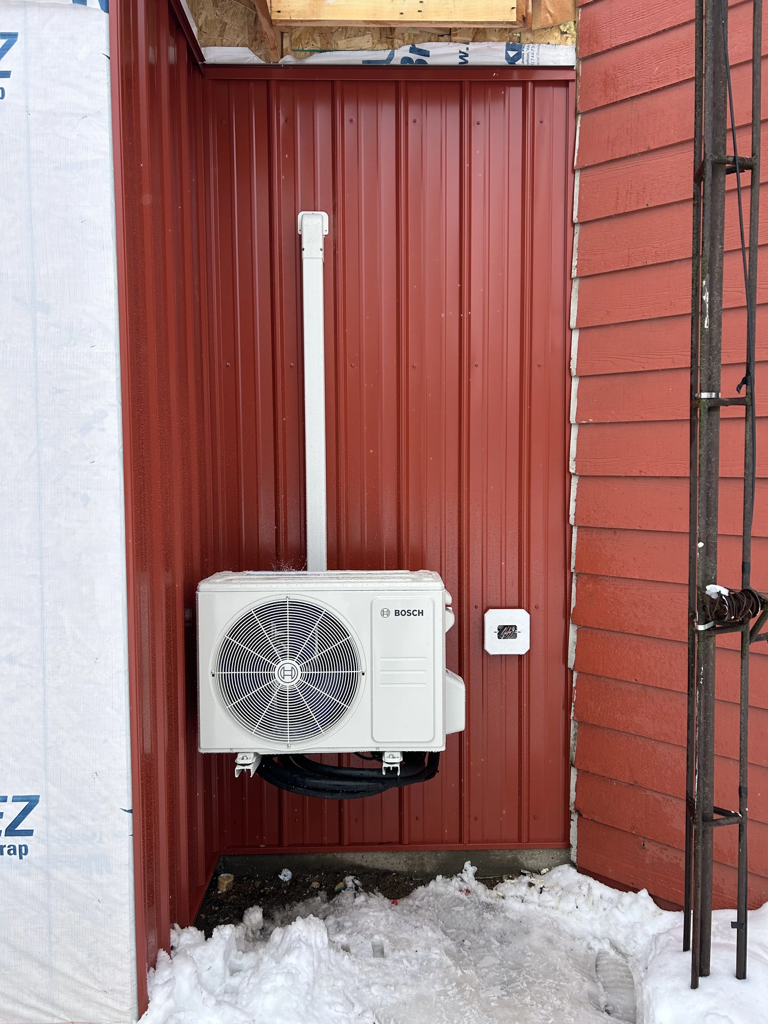 Outdoor heat pump system mounted on red metal siding with snow on the ground.