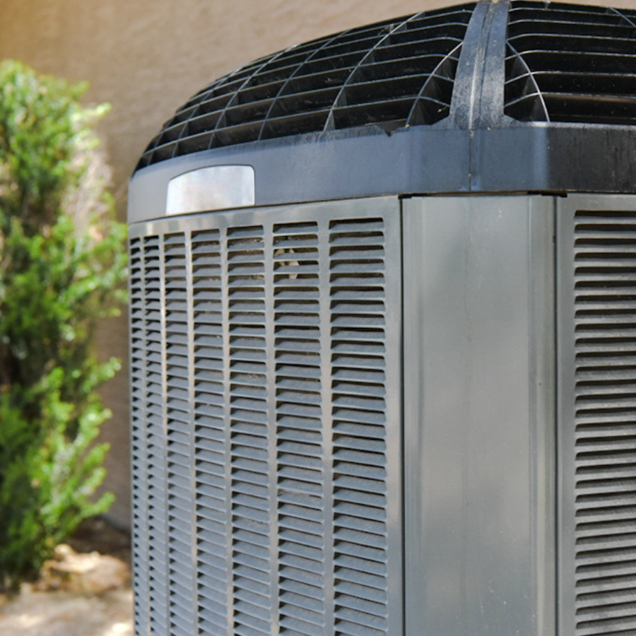 Close-up of an outdoor air conditioning unit with metal grilles and a fan on top, placed beside a wall and some green shrubbery.