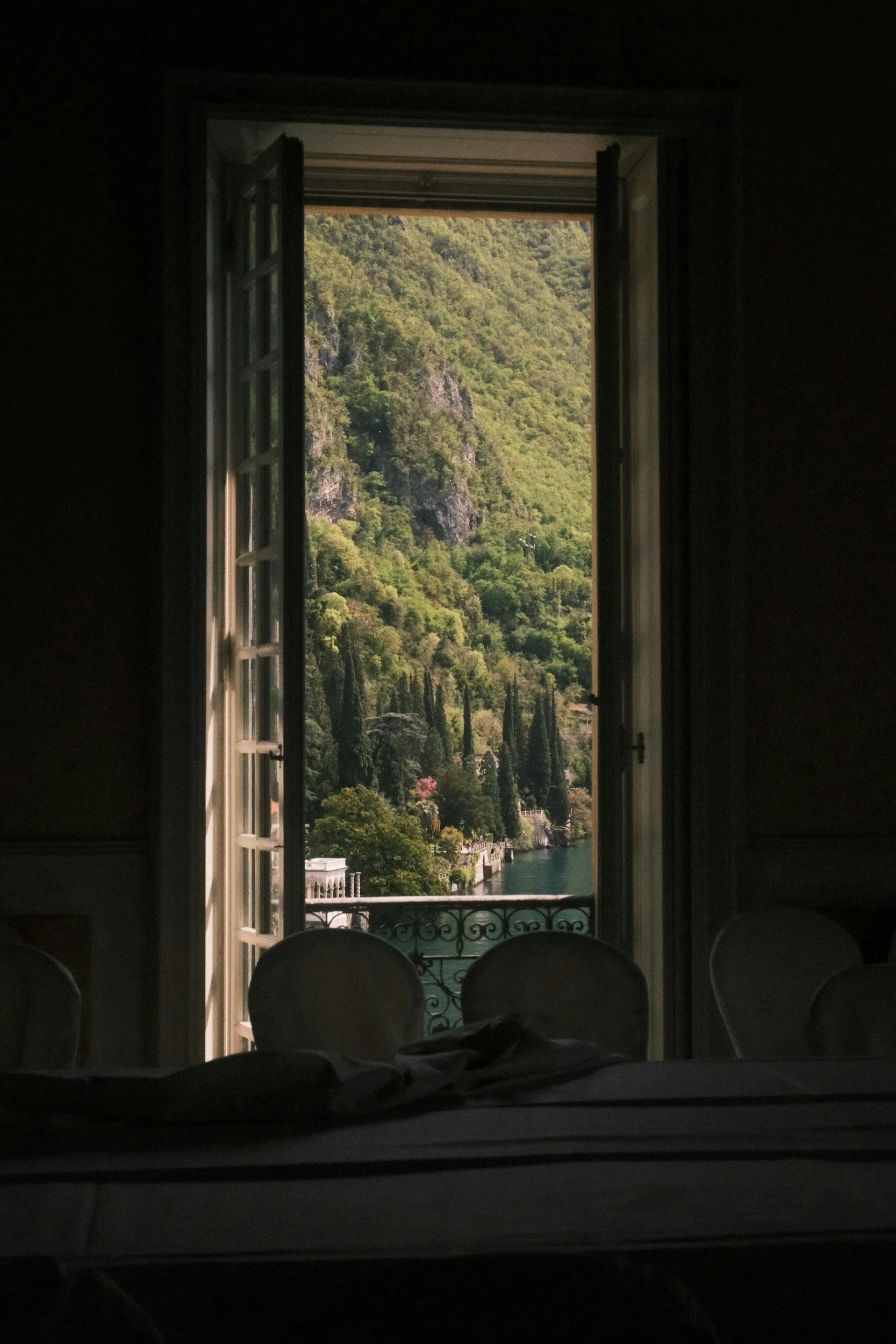 Open window overlooking lush green mountains, cypress trees, and a body of water with a balcony railing in the foreground.