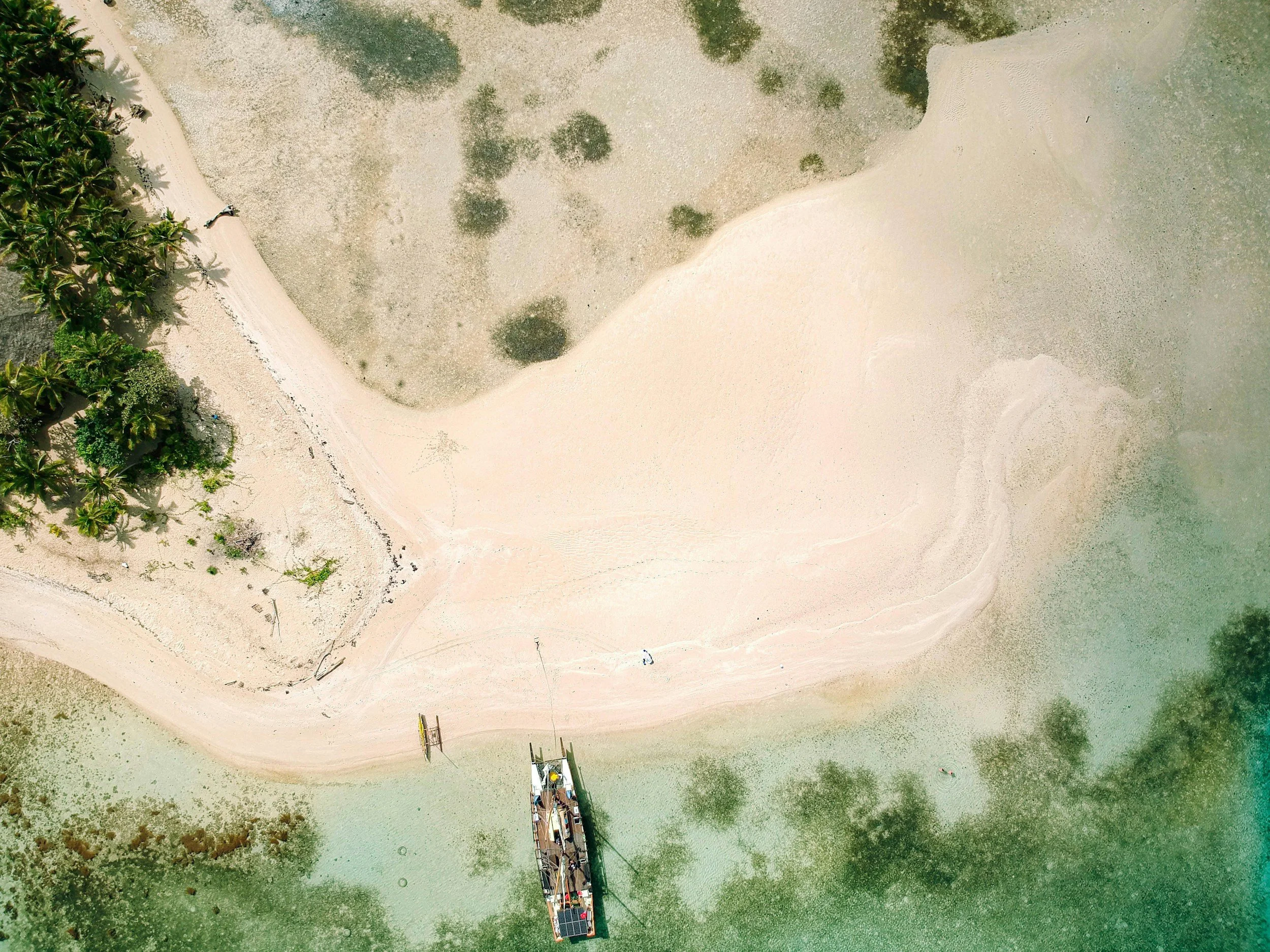 An aerial view of a small tropical beach with white sand, surrounded by clear green water, some trees, and a boat docked at the shore.