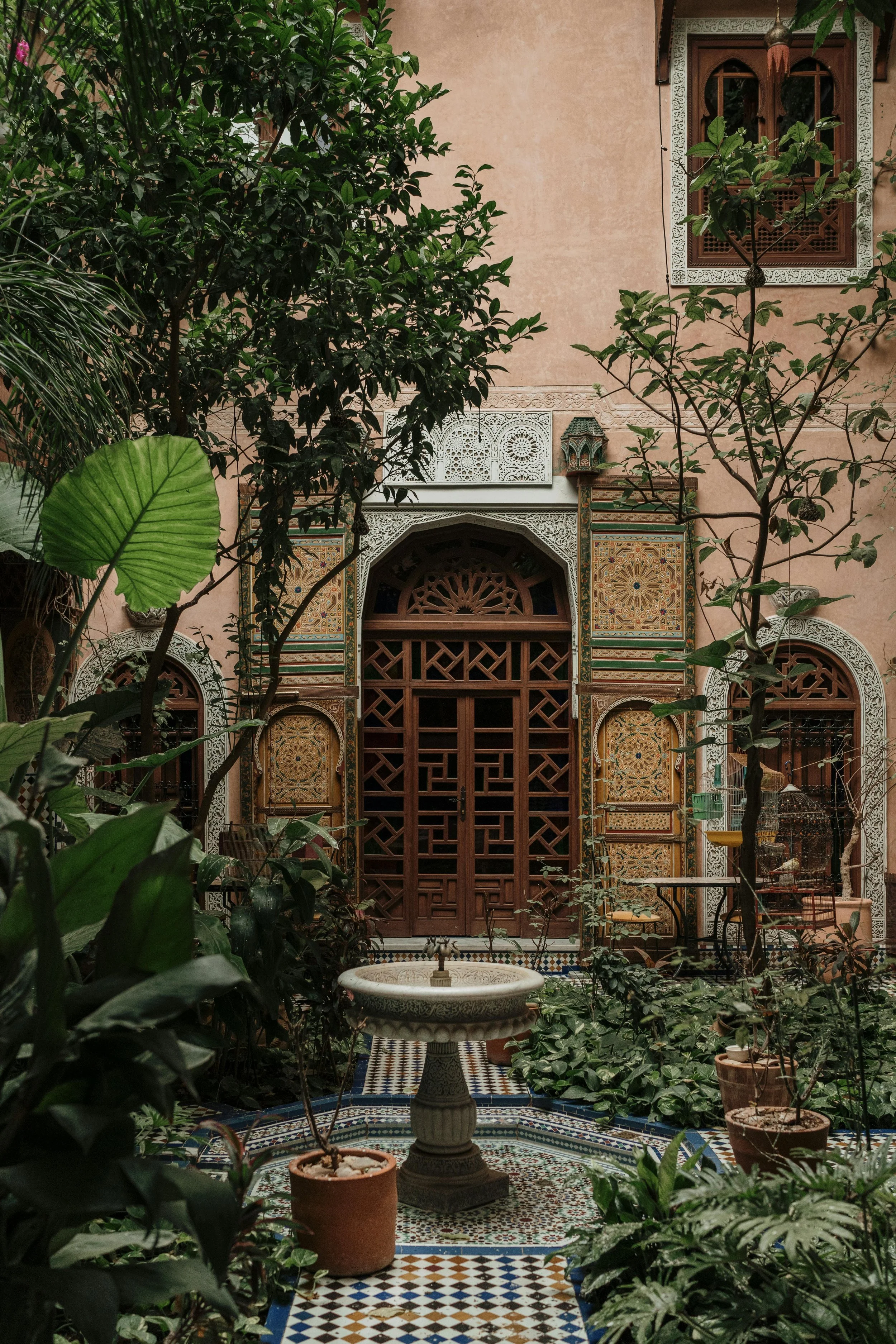 A lush courtyard with various green plants and a tiled floor. There is an ornate fountain in the center, surrounded by potted plants. The background features a detailed, decorative wall with stained glass windows, intricate woodwork, and Moroccan-inspired patterns.