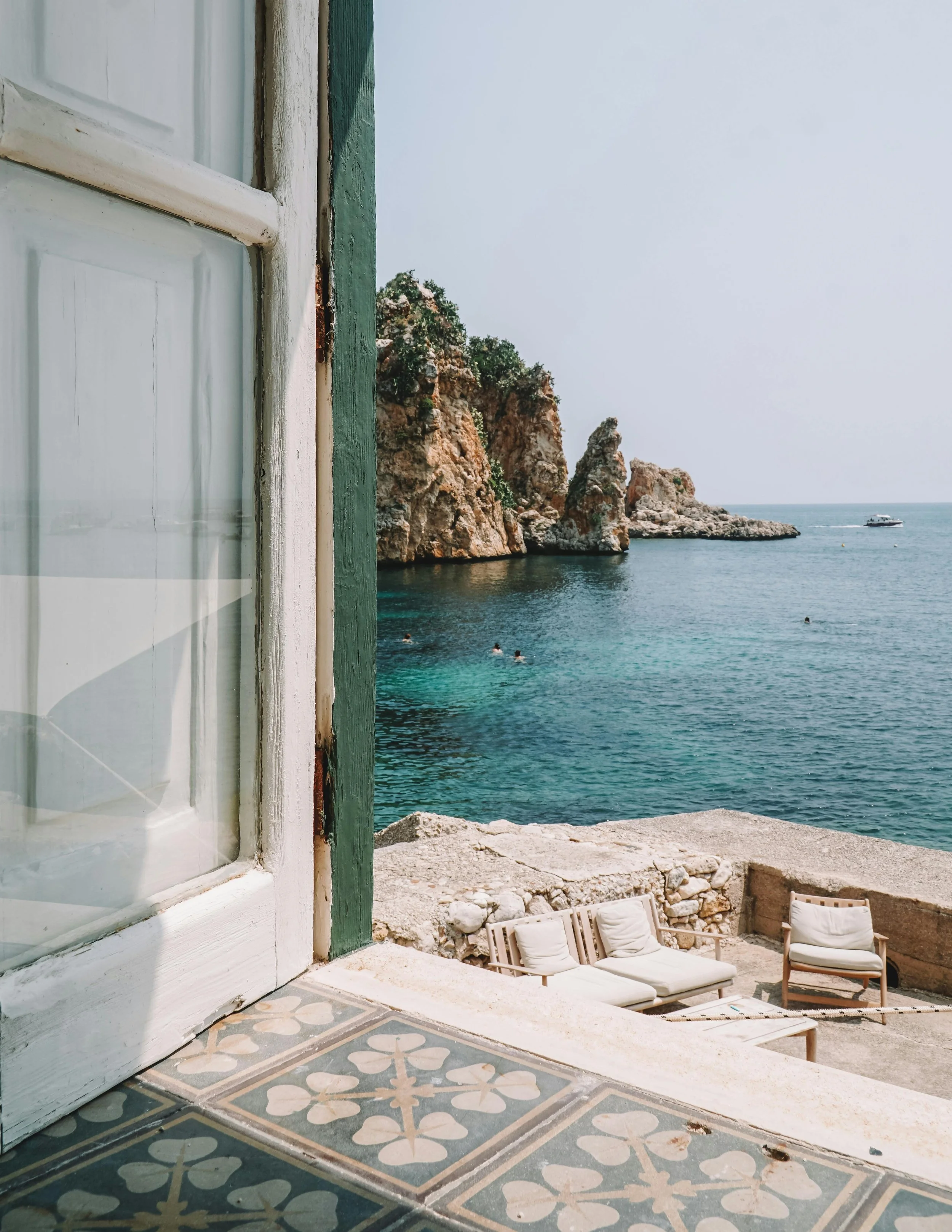 View of the sea through an open window, with rocky cliffs and boats in the distance, and outdoor furniture on a terrace.