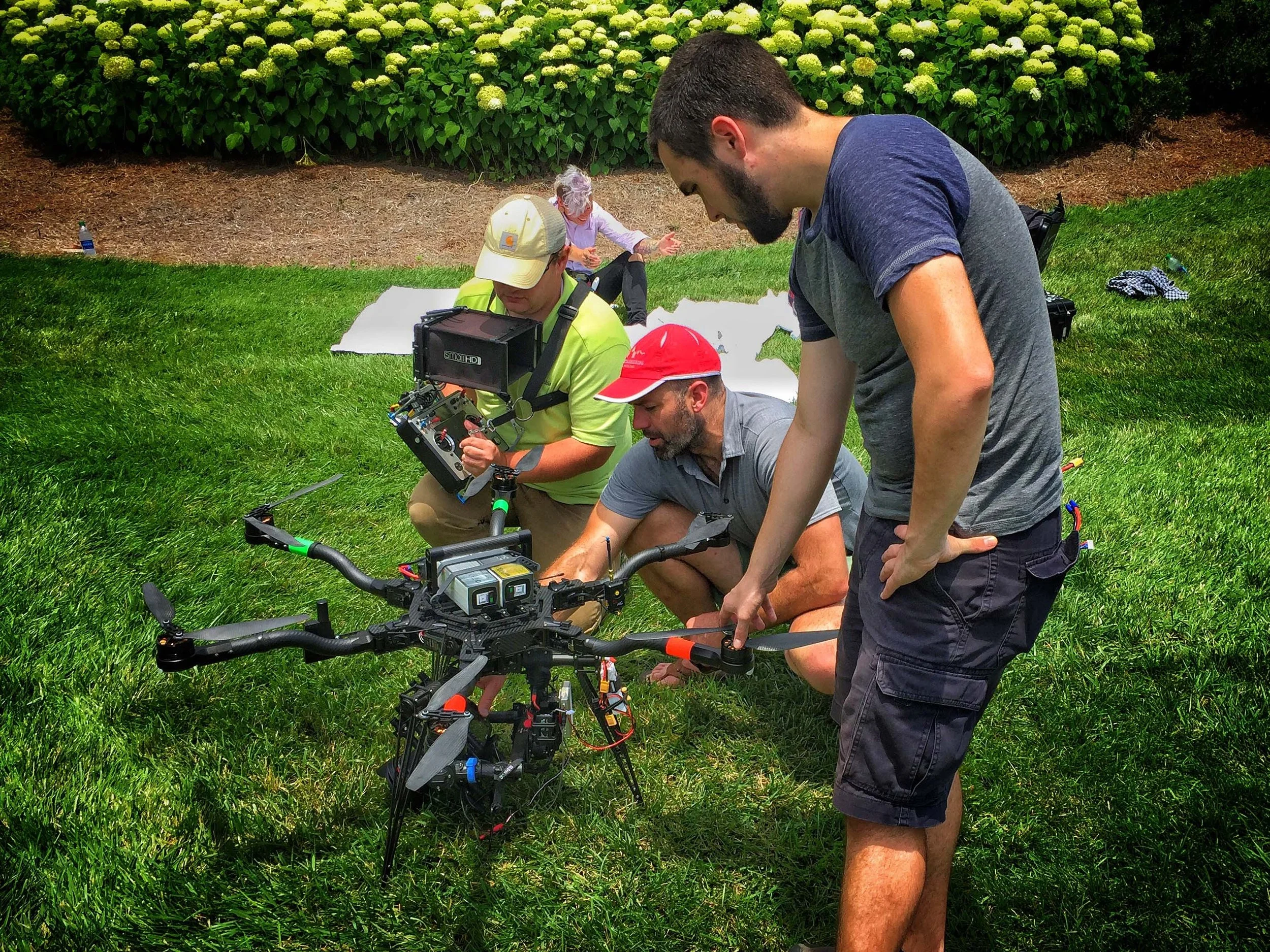 Group of three people working on a large drone in a grassy area, two men kneeling and one standing, with a woman sitting in the background on a blanket.