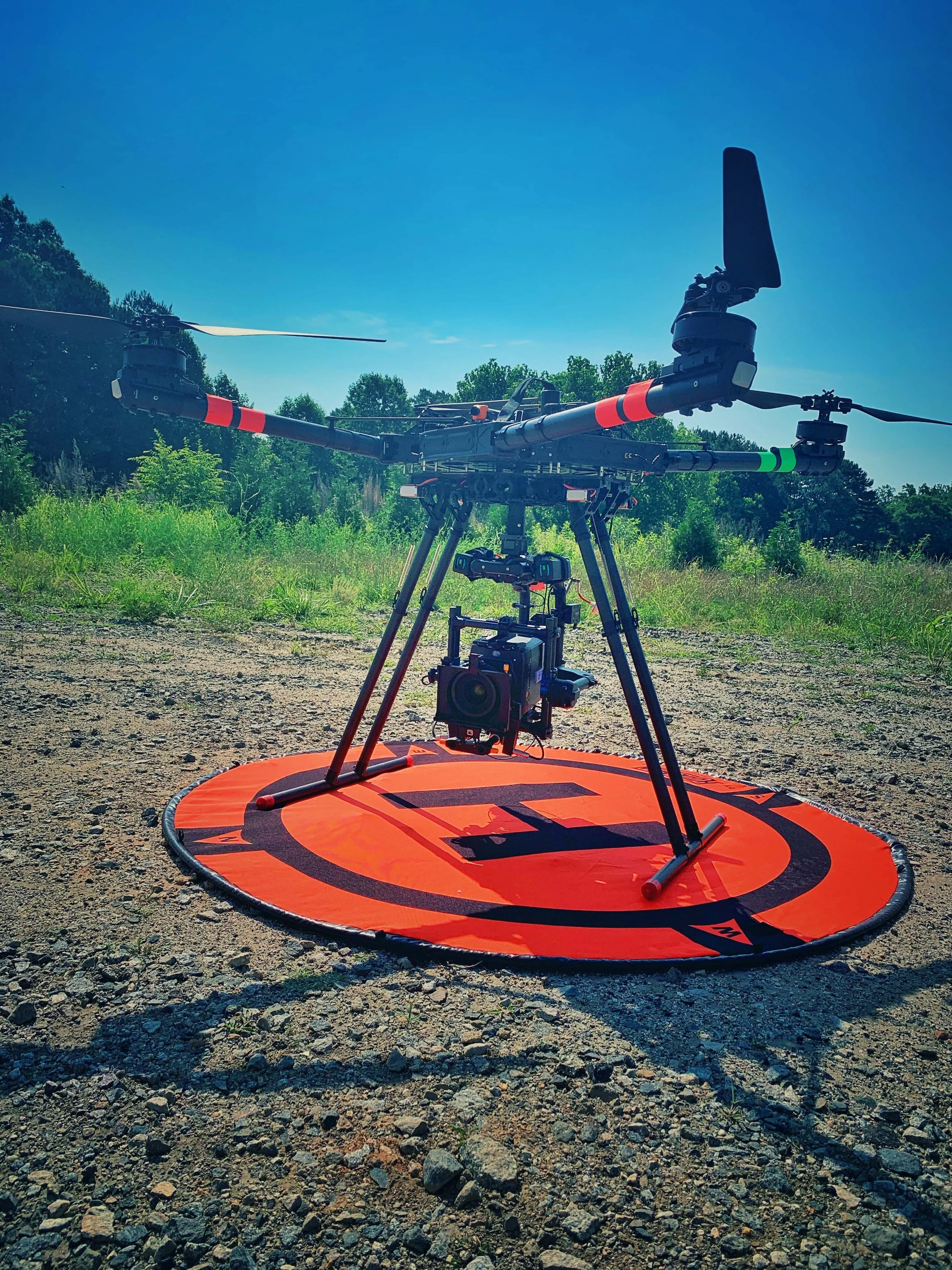 A large drone with multiple propellers on a bright orange landing pad outdoors, with trees and a blue sky in the background.
